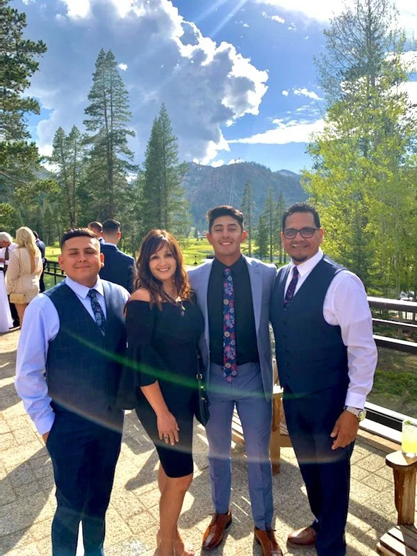 Four people in formal attire standing outdoors in front of a scenic mountain and forest background, smiling for a photo.