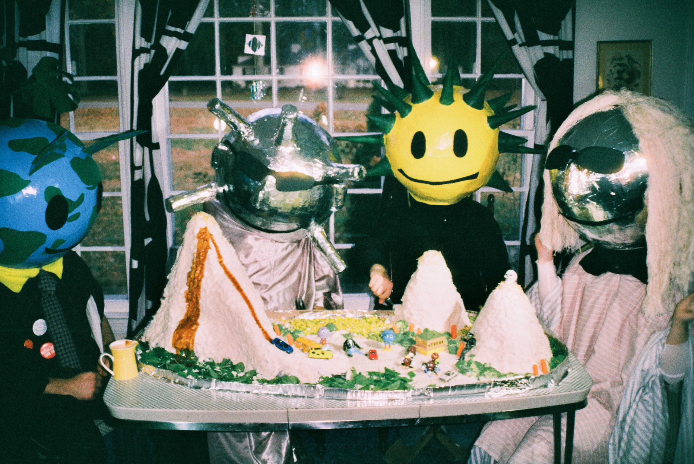 People wearing large, colorful, cartoonish masks around a cake at a party or celebration.