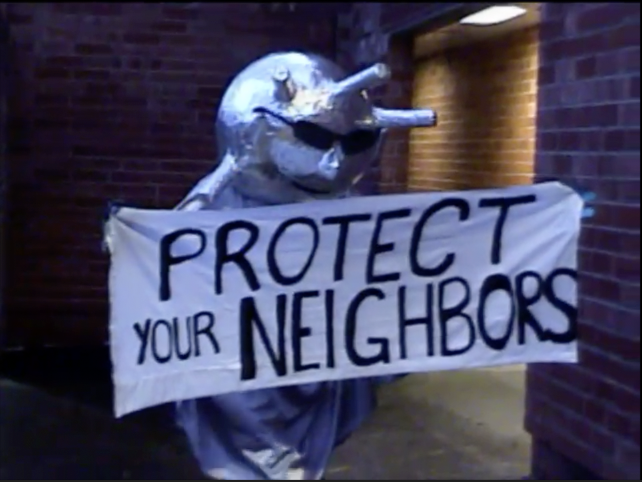 Person in a rhino costume holding a sign that says 'Protect Your Neighbors' outside a brick building at night.