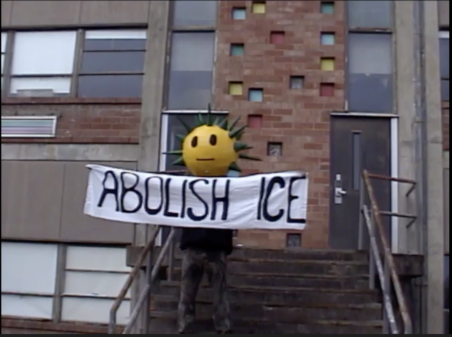 Person holding a protest banner that says 'Abolish ICE' on a staircase outside a brick building, with a large yellow smiling face mask with spiky green hair on their head.
