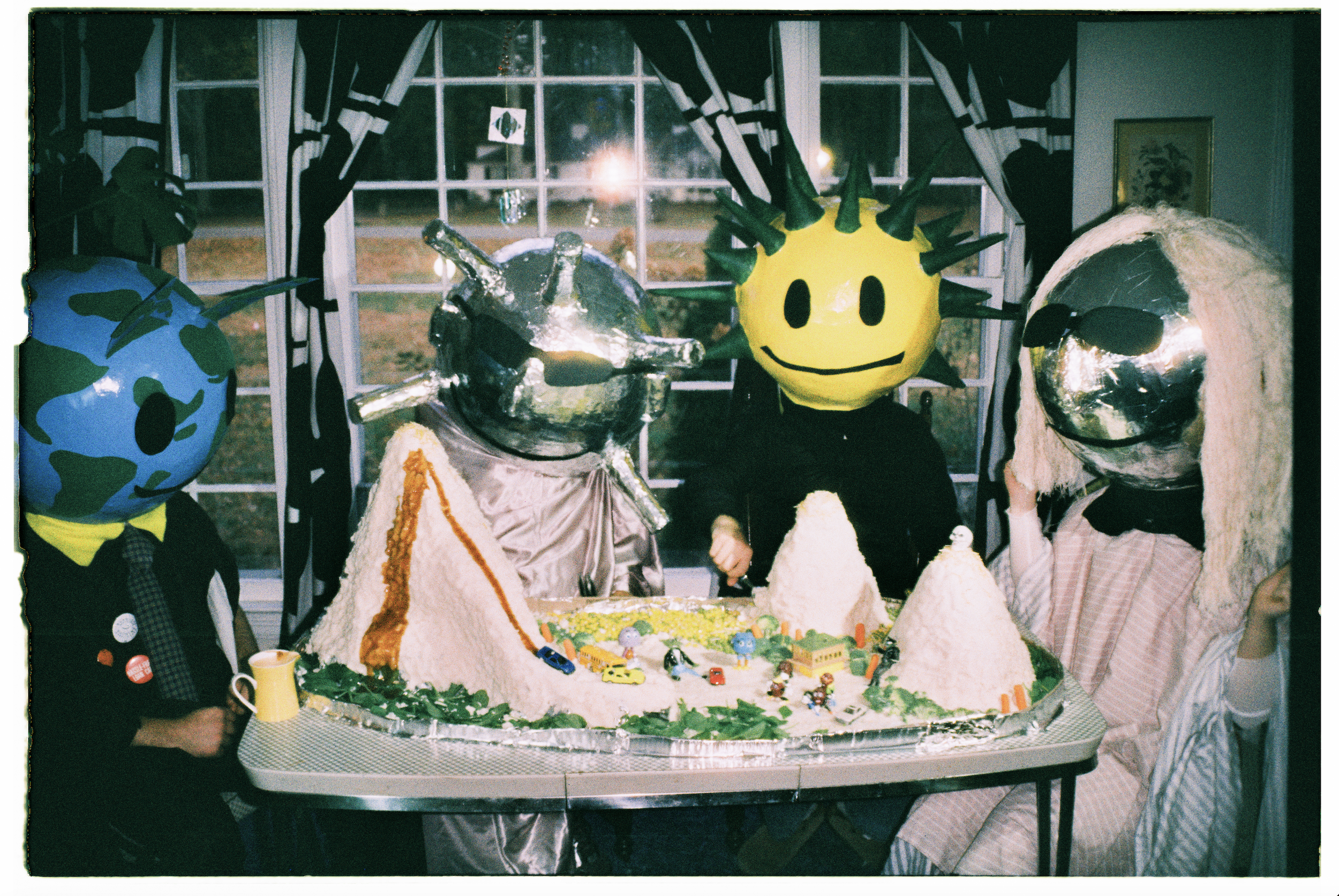 People wearing large, colorful masks with cartoon-like faces celebrating around a decorated birthday cake featuring mountains, roads, and toy cars inside a decorated room with a window backdrop.