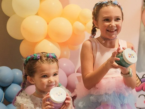 Two young girls with braided hair holding jars, at a birthday party with pastel balloons in the background.