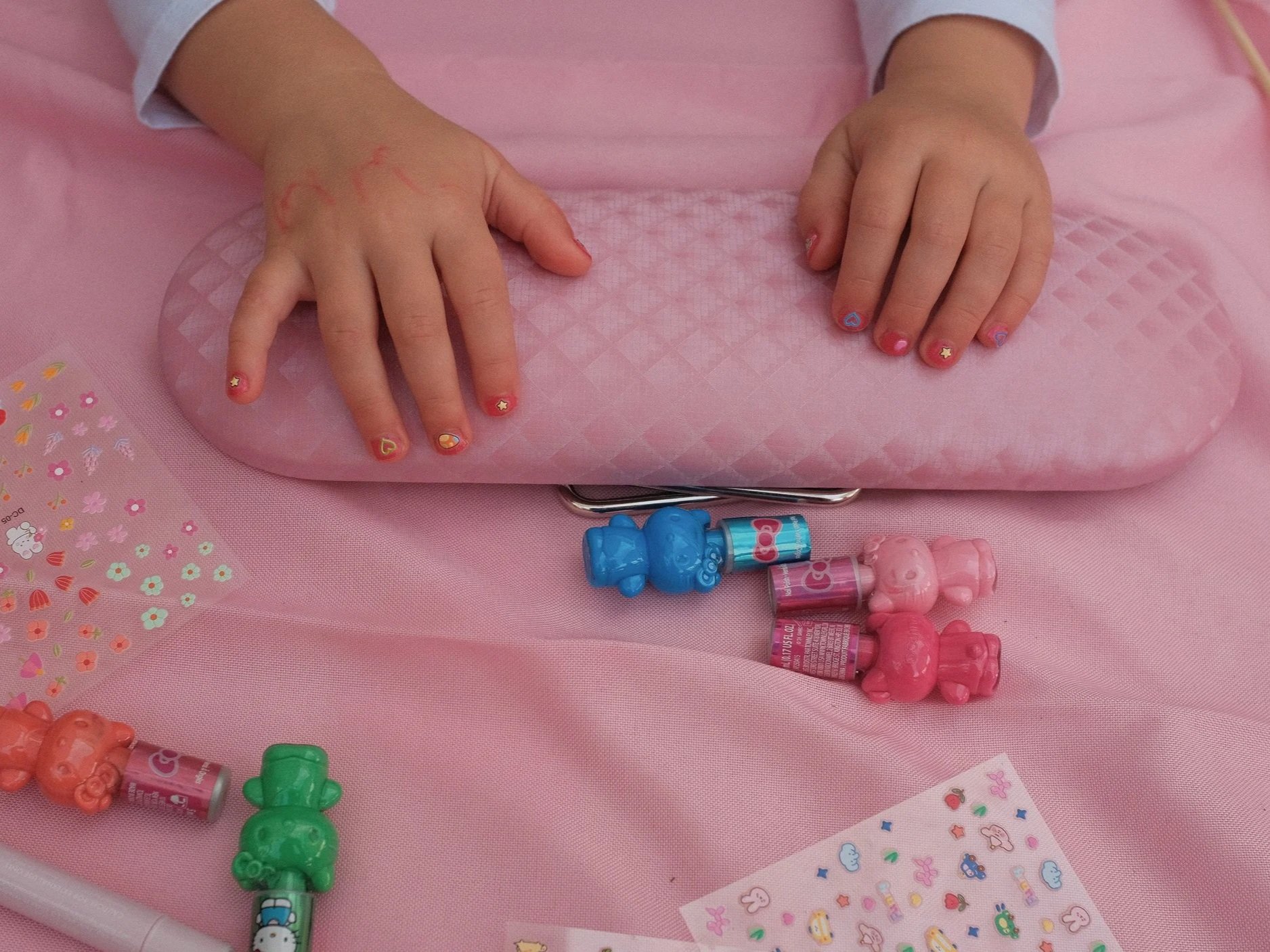 Close-up of a child's hands with colorful nail art, resting on a pink cushioned surface surrounded by small decorative stickers, teddy bear-shaped lip balms, and other small toys on a pink tablecloth.