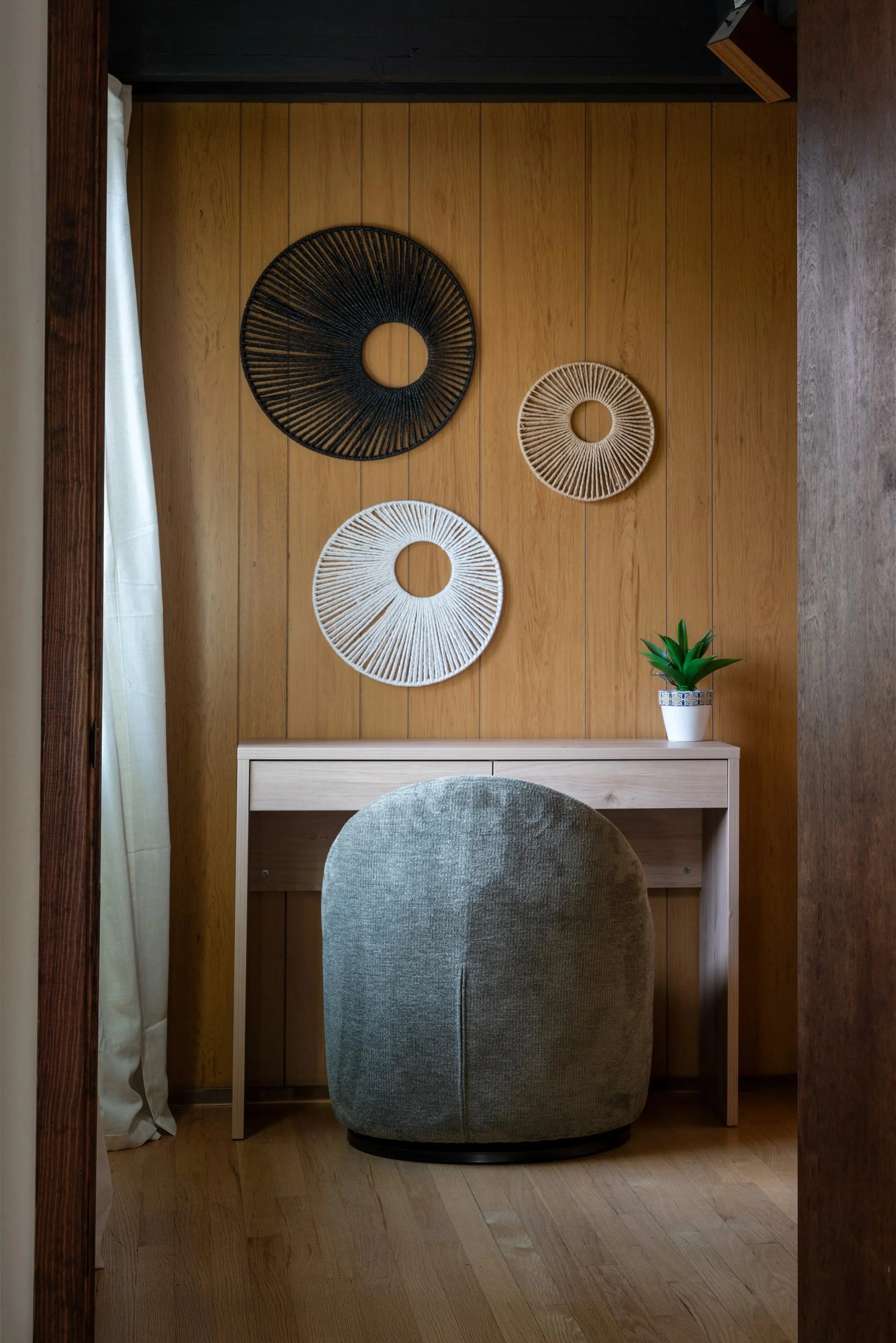 A small workspace with a wooden wall, a white desk, a grey upholstered chair, a small potted plant, and decorative circular wall hangings.