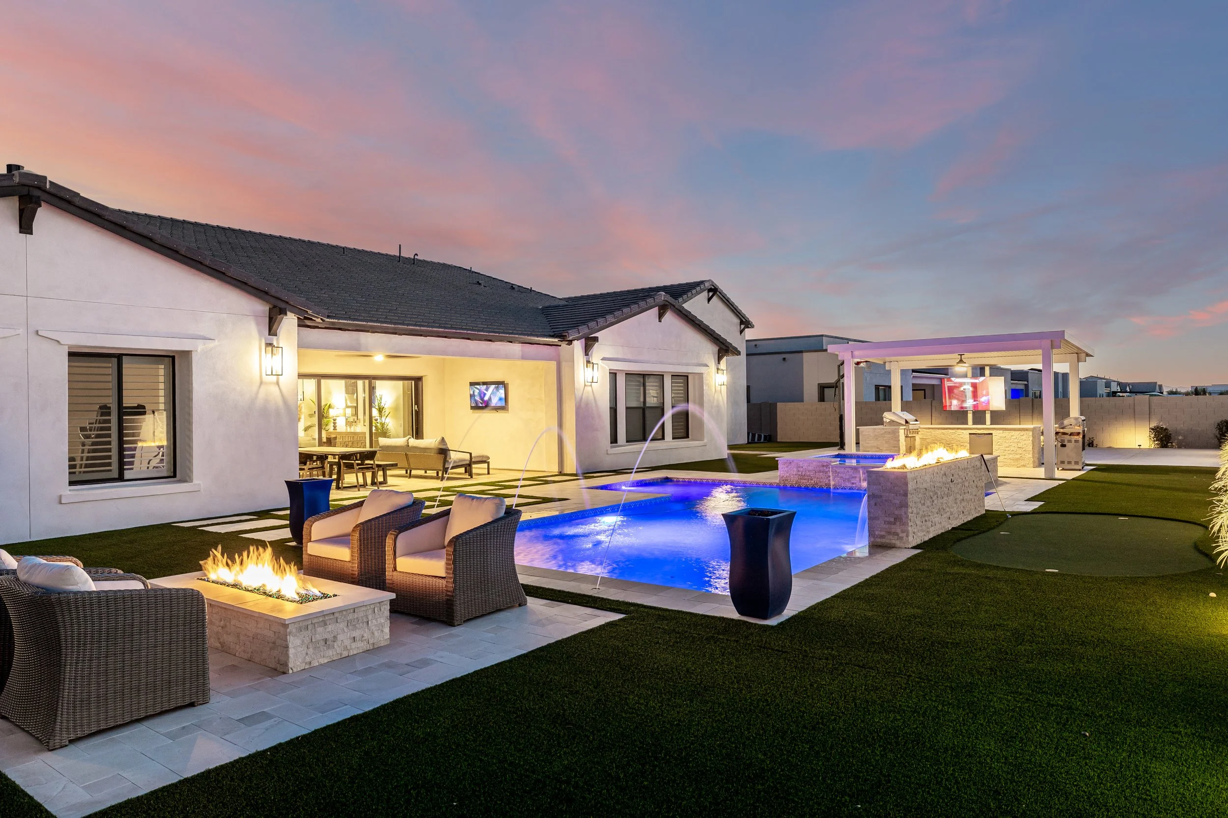A backyard with a swimming pool, outdoor seating, fire pits, and a covered patio area at sunset, surrounded by a lawn and modern house.