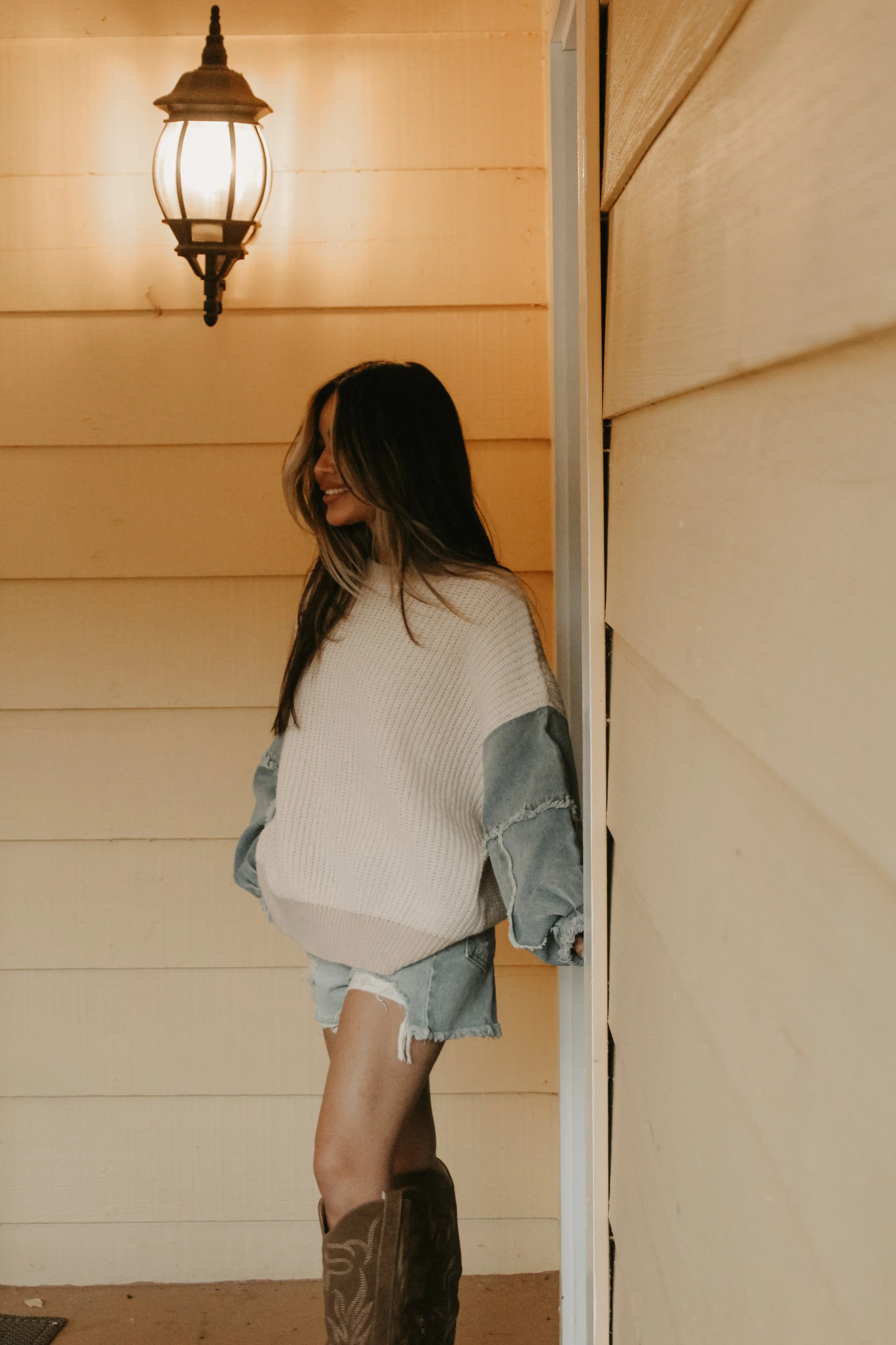 A woman with long hair and a smile, wearing a white sweater with denim accents on the sleeves, ripped denim shorts, and cowboy boots, standing outside a house with light-colored wooden siding and a wall-mounted lantern.