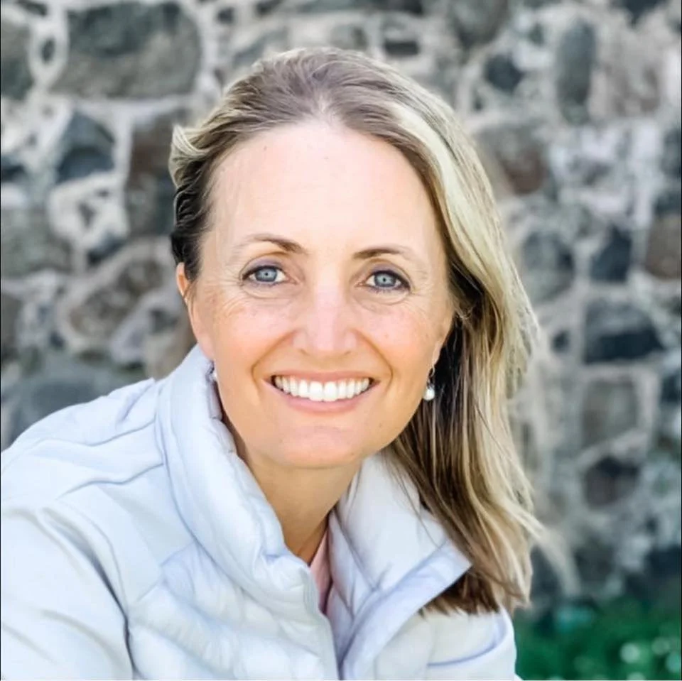 A woman with blonde hair, blue eyes, and a bright smile, wearing a white jacket, outdoors with a stone wall in the background.