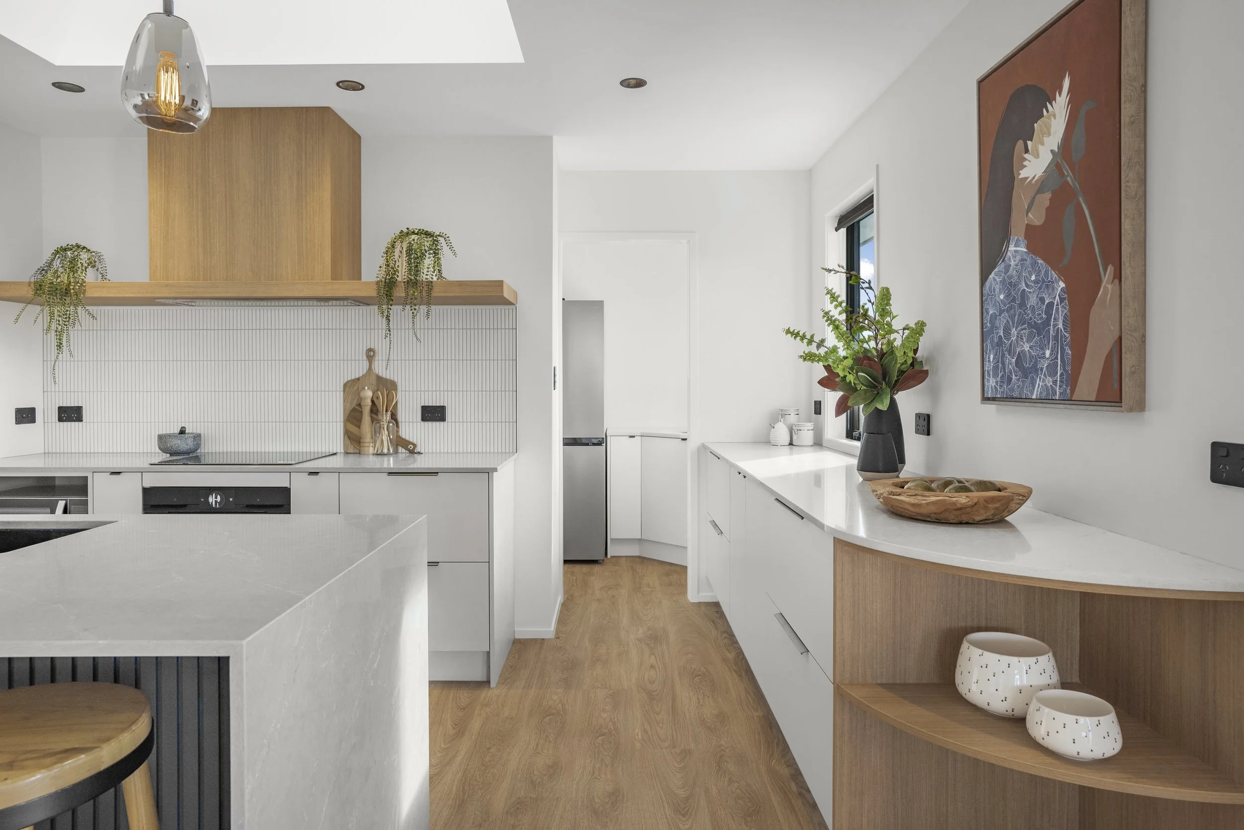 Modern kitchen with white cabinets, wooden accents, and a wooden floor. Decor includes a vase with greenery, a framed artwork of a woman, and various bowls and jars.