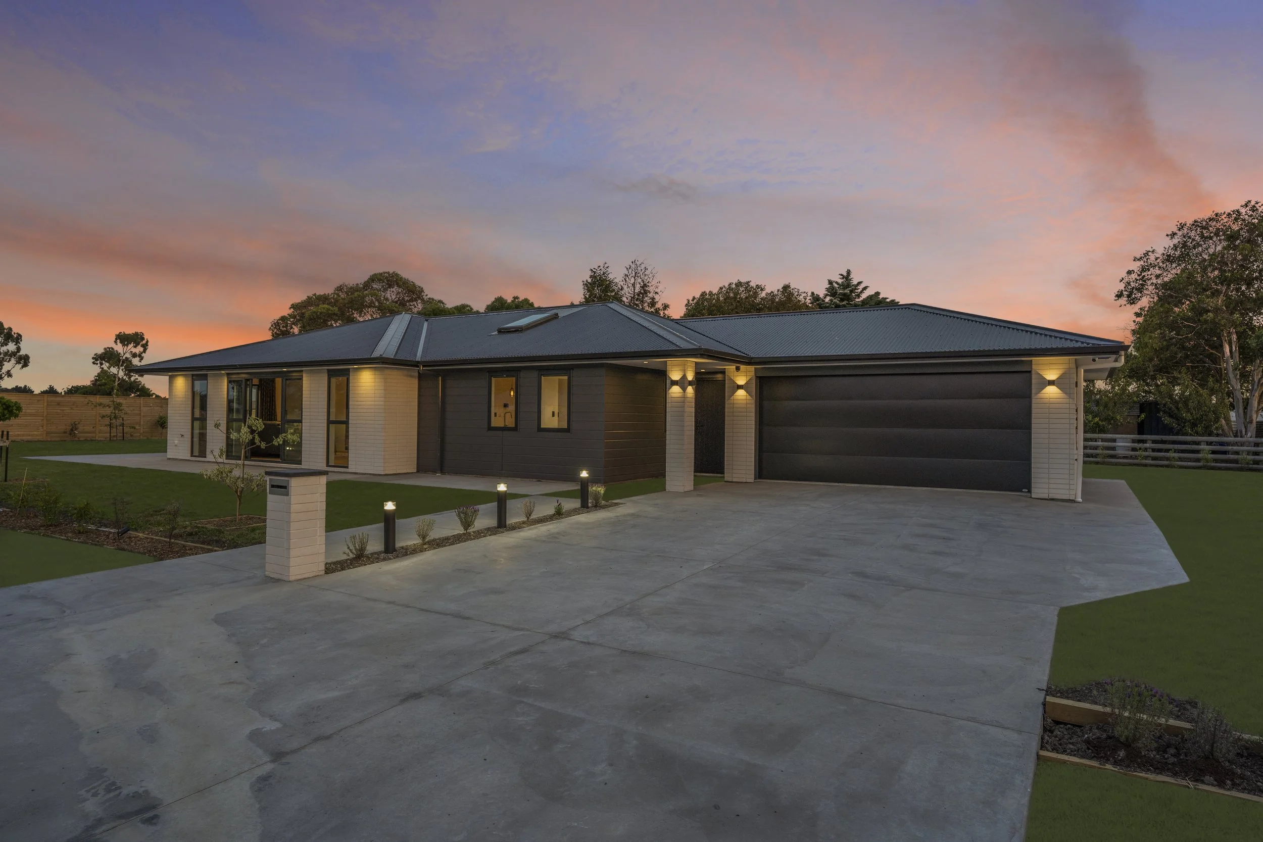 Modern single-story house with a dark grey roof and a combination of dark grey and white exterior walls, illuminated at dusk with a driveway, small landscaped lawn, and trees in the background.