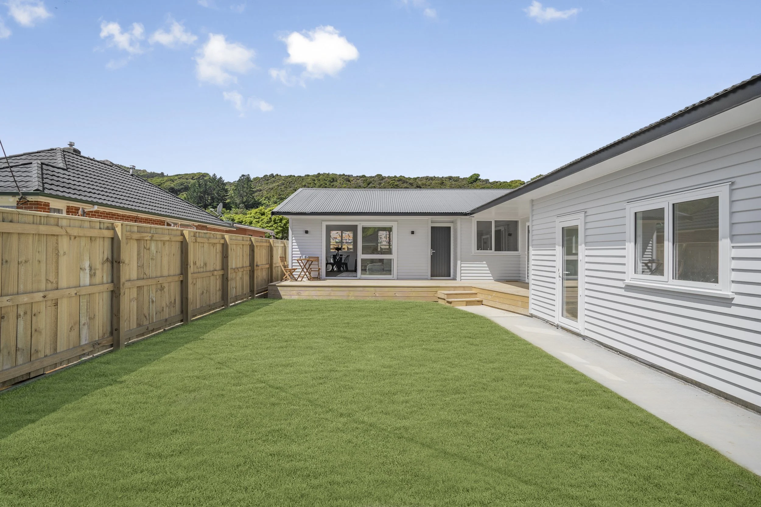 A backyard with green grass, a wooden fence on the left, a white house with siding on the right, a patio with wooden steps leading to a deck with outdoor furniture, and a clear blue sky with a few clouds.