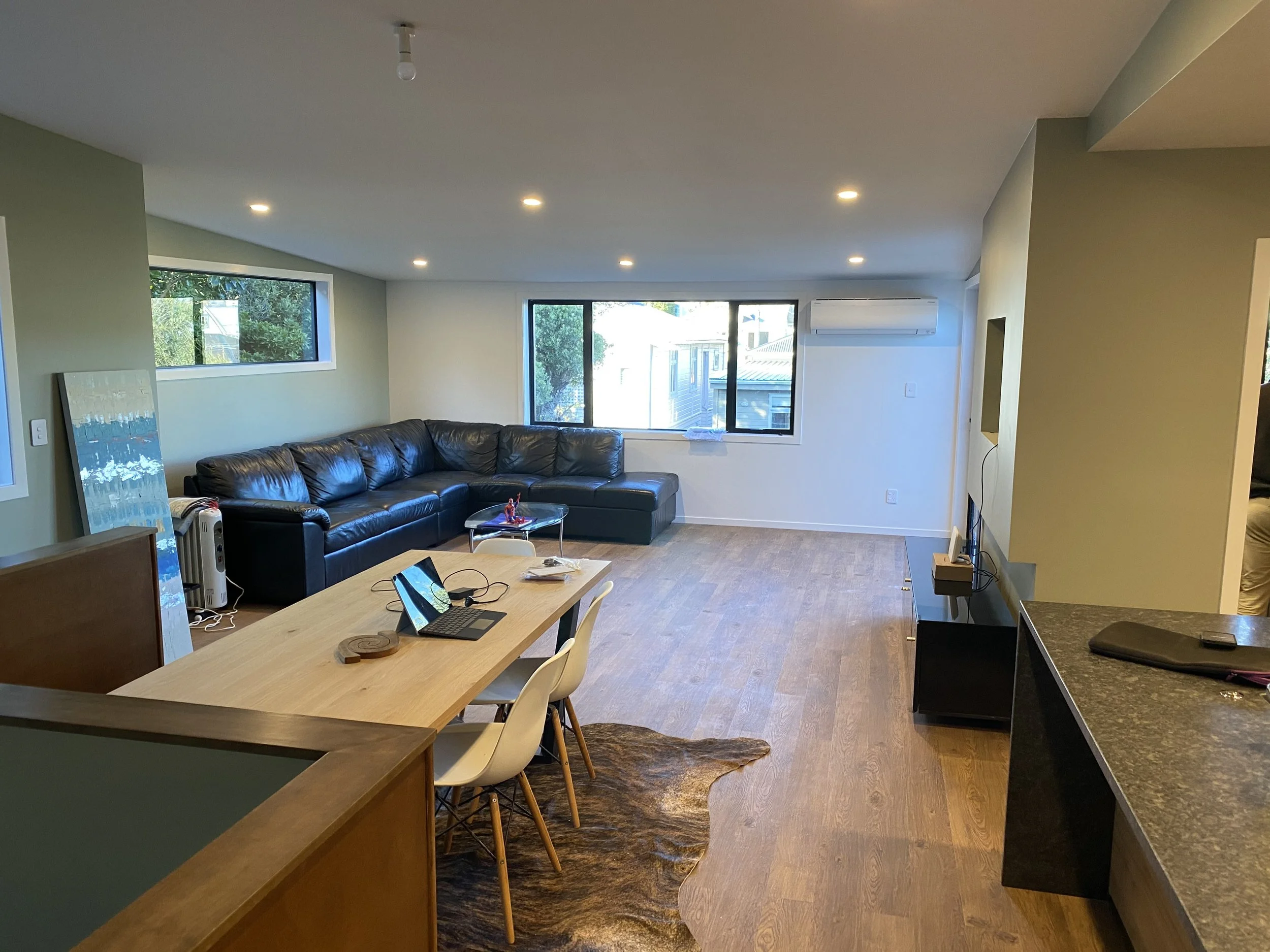 Interior of a living room with a black leather sectional sofa, large windows, wooden flooring, and a dining table with chairs.