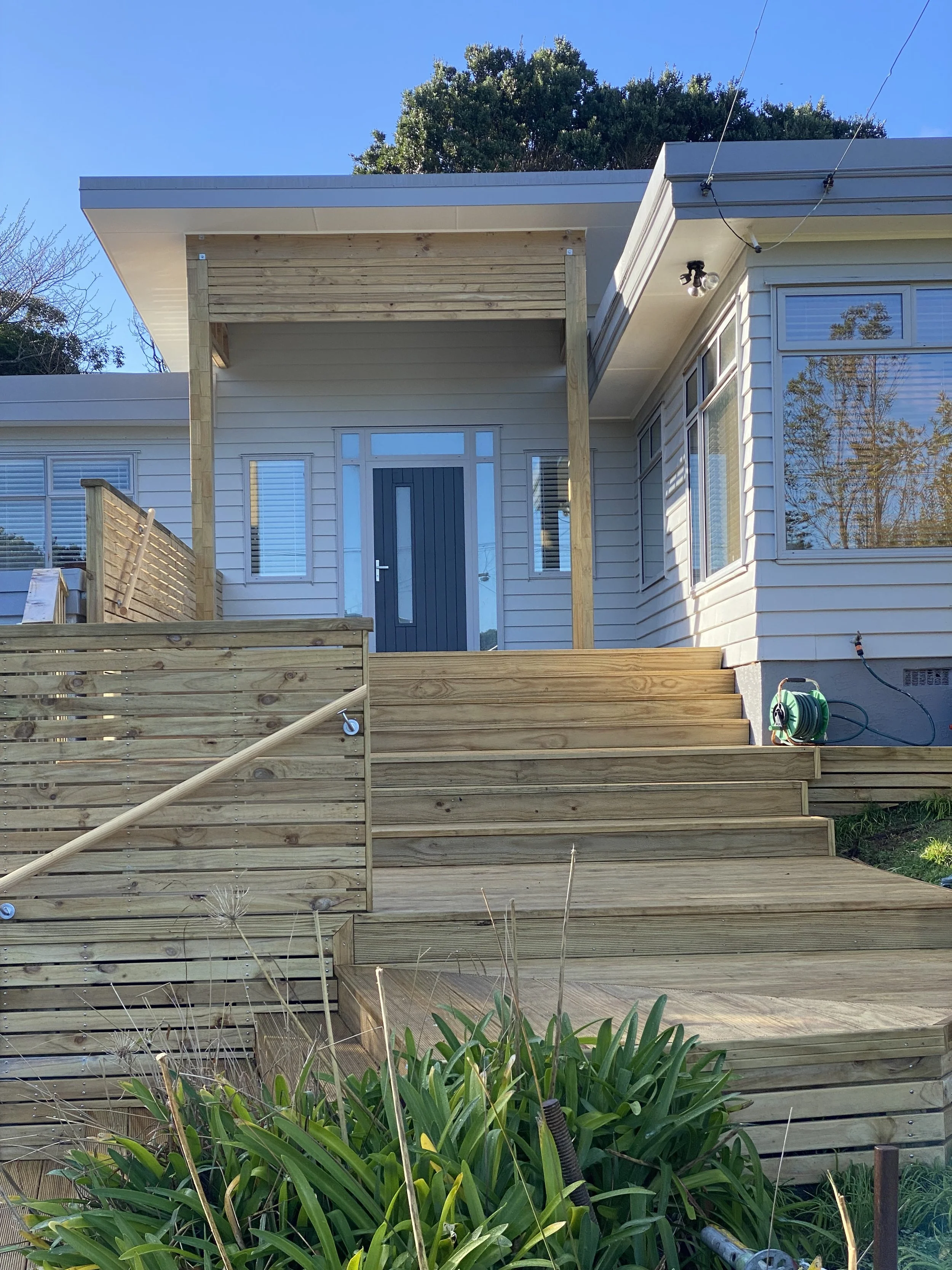 Front view of a house with a new wooden deck, stairs, and porch support beams, with a garden in the foreground and blue sky above.