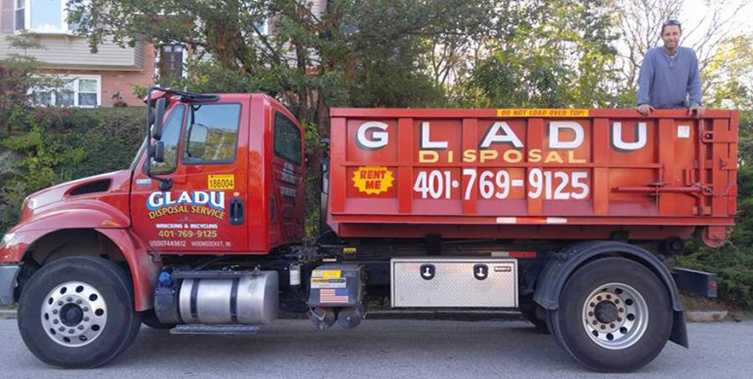 A red disposal truck for Glad U Disposal, with a man standing on top, parked on a street in front of greenery and residential buildings.