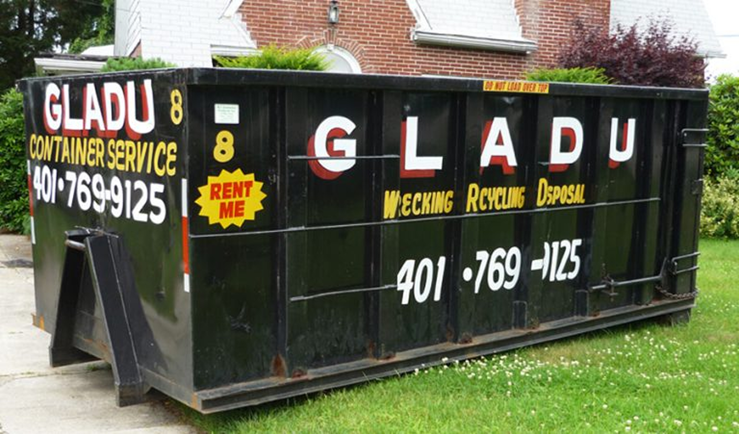 Black dumpster with white and red lettering advertising GladU container service, located on a grassy yard with houses and bushes in the background.
