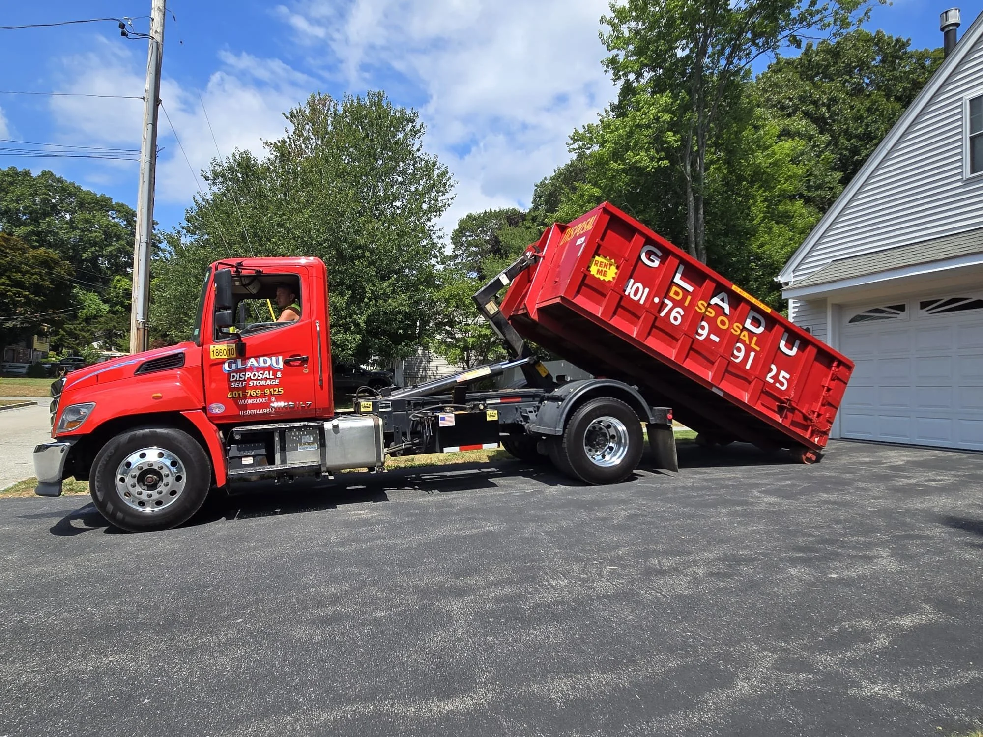 A red dumpster truck parked on a driveway with the dumpster slightly elevated at an angle. The truck has company branding, including a phone number, and is set in a residential neighborhood with trees, houses, and a partly cloudy sky in the background.