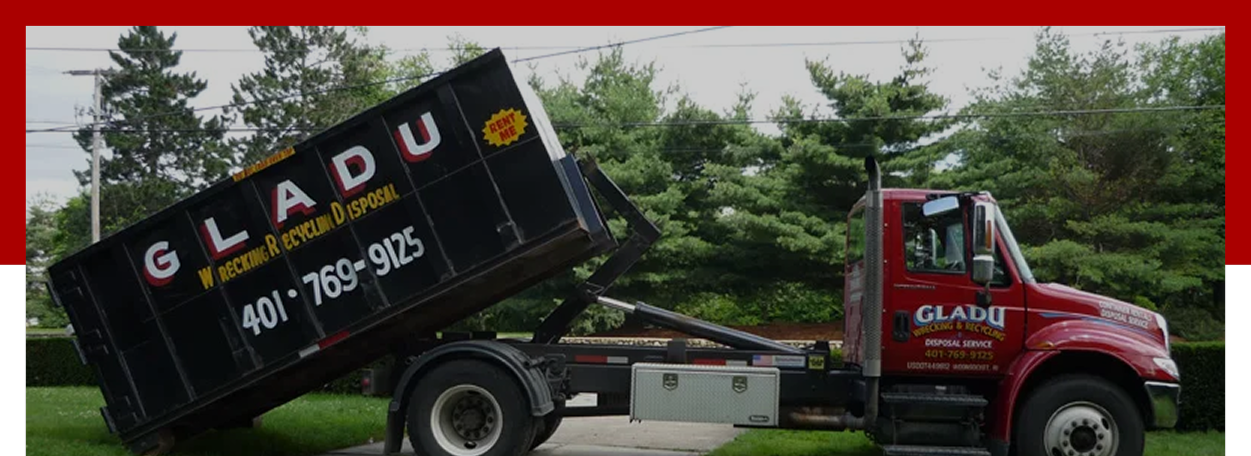 A red garbage truck tipping a black waste container with yellow and white lettering, parked on a grassy area with green trees and power lines in the background.