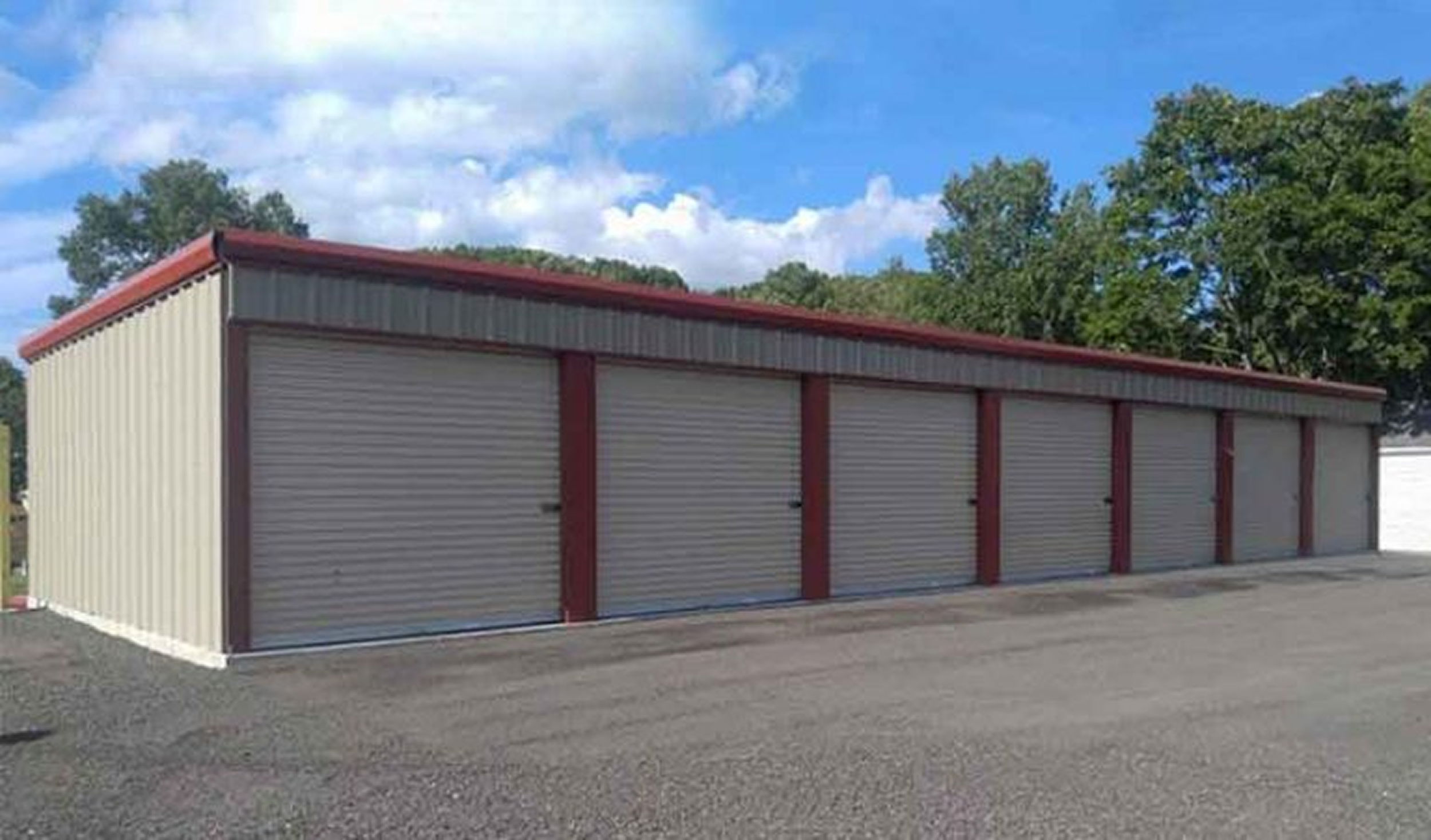 A beige storage unit with multiple closed metal roll-up doors, red trim on the edges, situated on a gravel lot with trees and blue sky in the background.