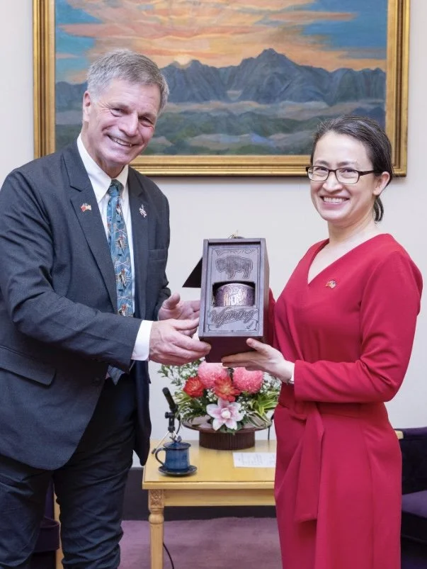 A man and a woman smiling as they exchange a gift in a room with a landscape painting on the wall, a table with a flower arrangement, and a purple chair.