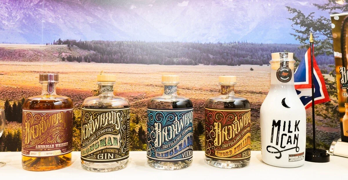 Five bottles of flavored whiskey from the Hawkridge Distilling Company lined up on a table with a scenic mountain backdrop. The bottles each have distinct, colorful labels, and the second bottle from the right features a small British flag and a milk bottle with the text "Milk Can."