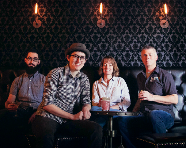 Four people sitting on a black leather booth in a dimly lit bar with black damask wallpaper and wall-mounted candles.