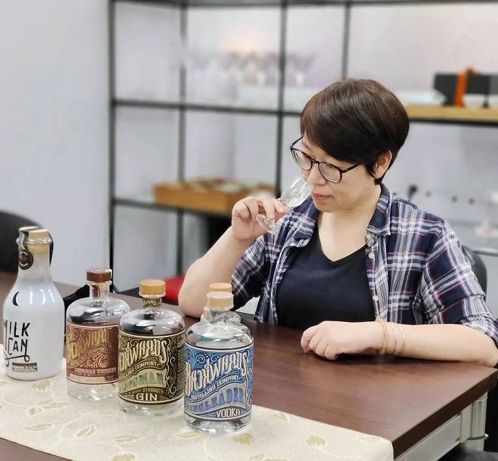 A woman with short dark hair and glasses tasting a bottle of vodka, surrounded by other bottles of liquor on a table in a modern room.