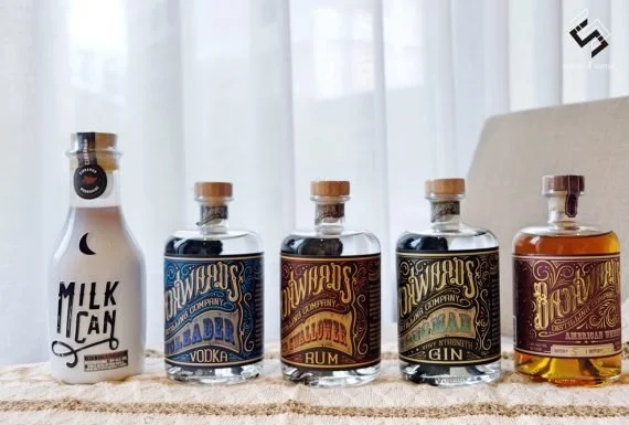 Collection of five miniature bottles of alcohol and a milk carton shaped bottle on a table, with a curtain background.