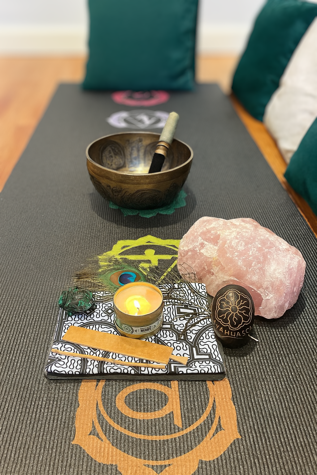 A table with a pink Himalayan salt rock, a candle, a peacock feather, a small green stone, and a black and gold decorative object, all on a patterned cloth. In the background, there is a singing bowl with a mallet and colorful cushions.