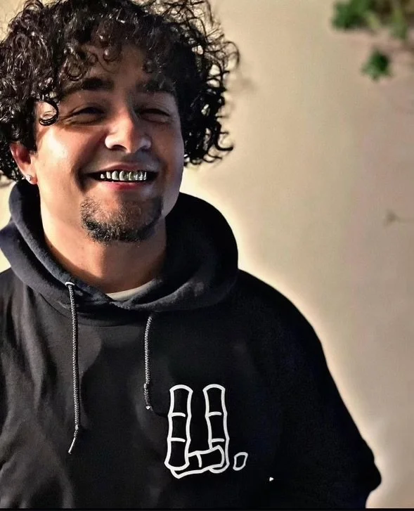 A man with curly hair smiling, wearing a black hoodie with a white hand making a rock on sign on it.