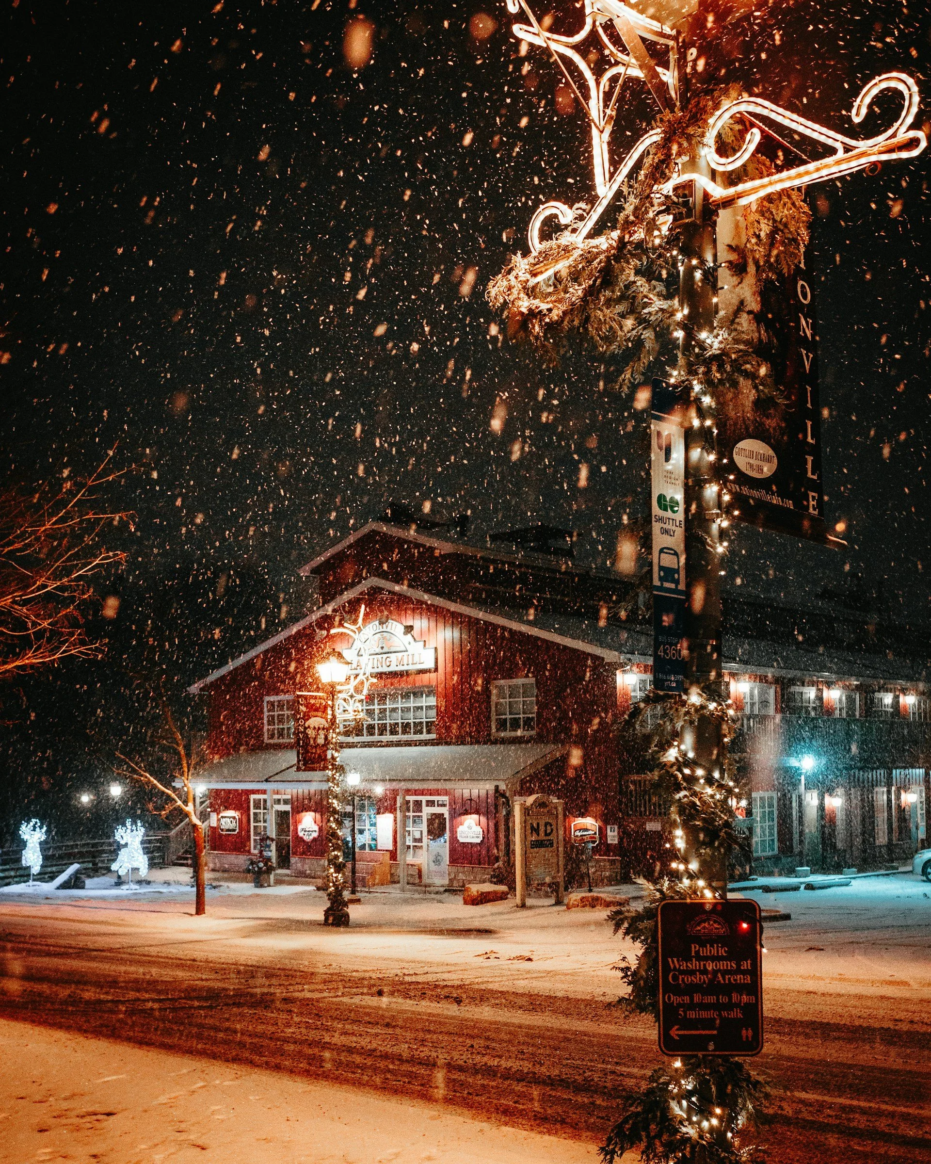 Snow falling at night outside a decorated building with bright lights and Christmas decorations. There are snow-covered trees and a street sign indicating public washrooms and a walking distance to Crosby Arena.