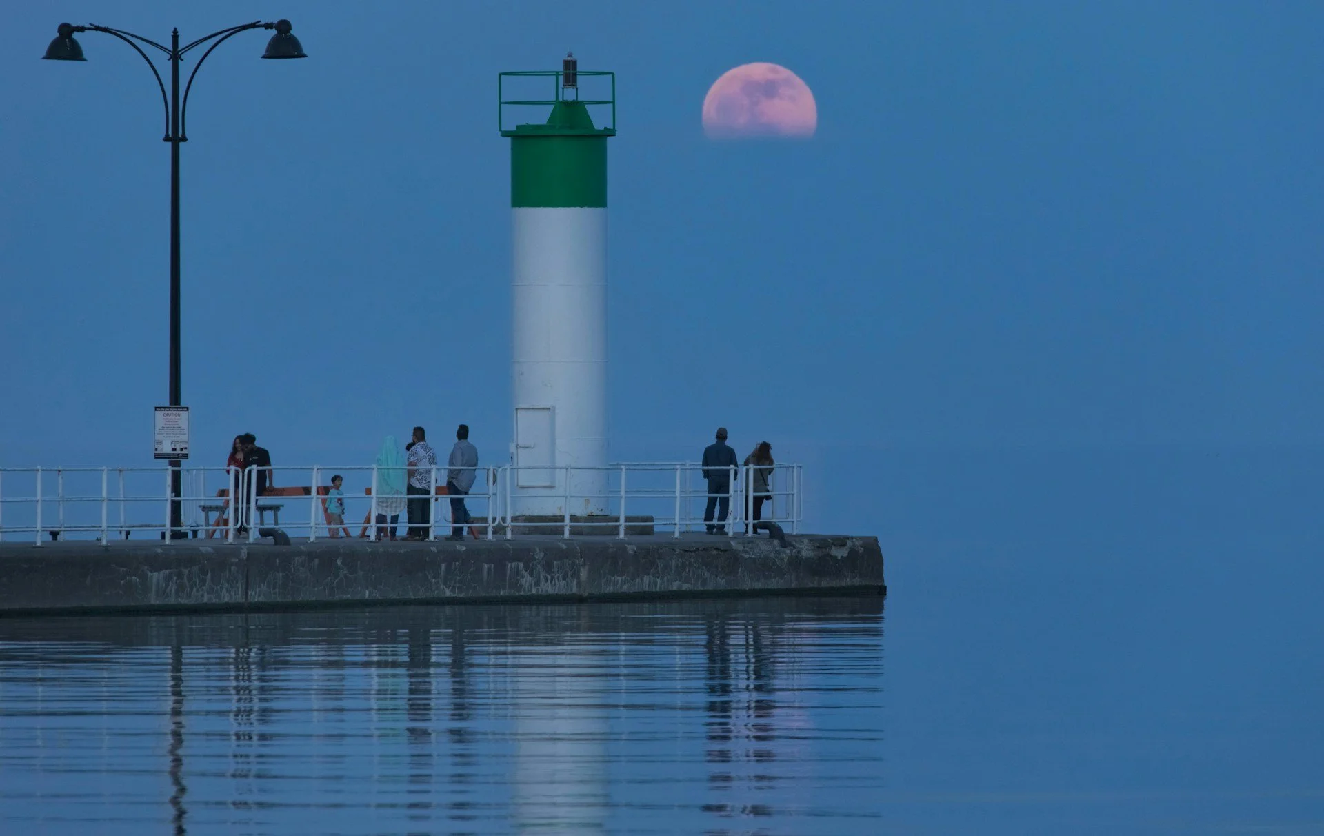 A group of people standing on a pier with a green and white lighthouse, moon rising in the background, and calm water reflecting the scene.