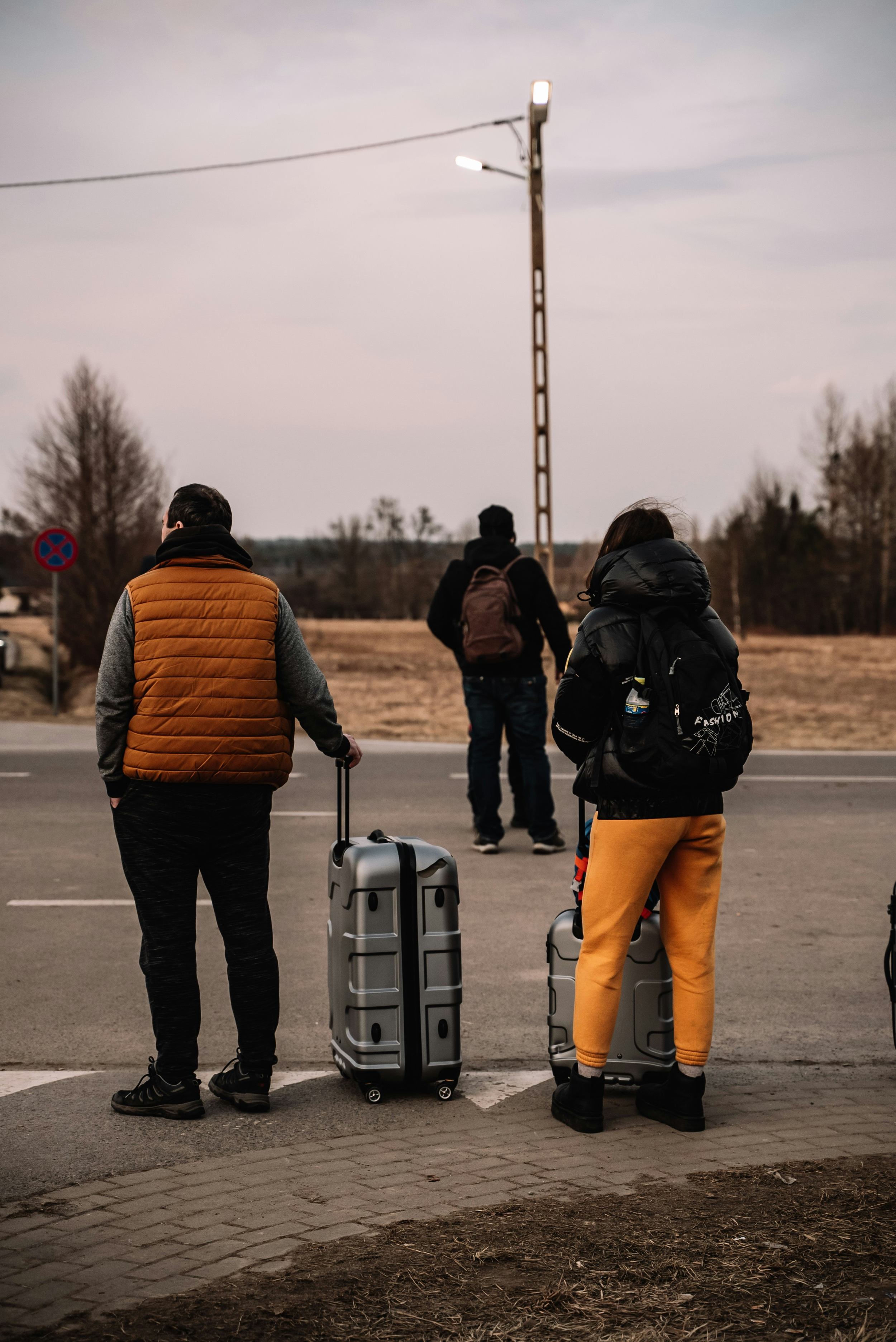 Three newcomers waiting with luggage at an outdoor transit area, representing travel and the immigrant journey to Canada.