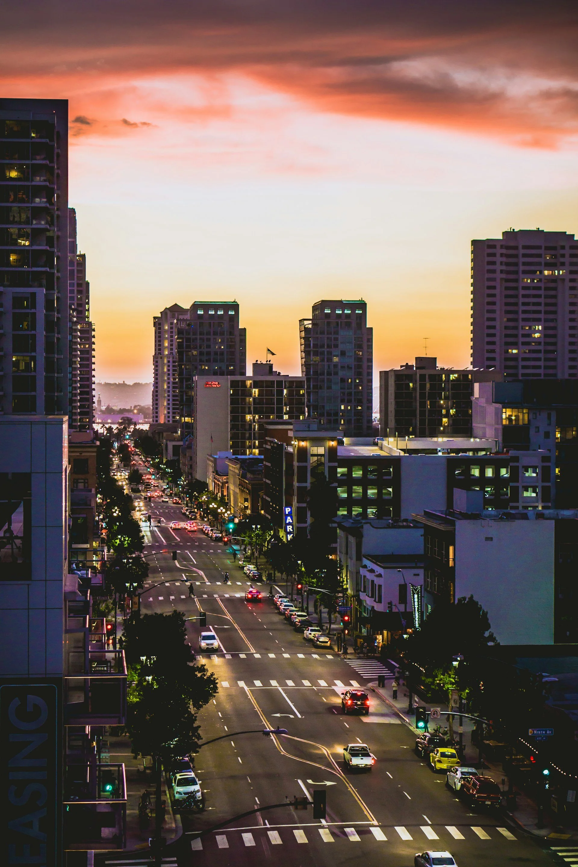 Cityscape at sunset with tall buildings, a busy street with cars, and a colorful sky with pink and orange clouds.