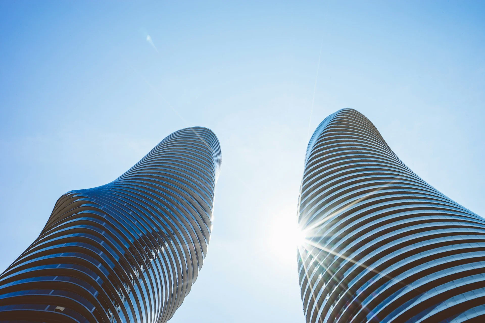 Low-angle view of two modern skyscrapers with curved, striped facades against a bright blue sky with sun glare.