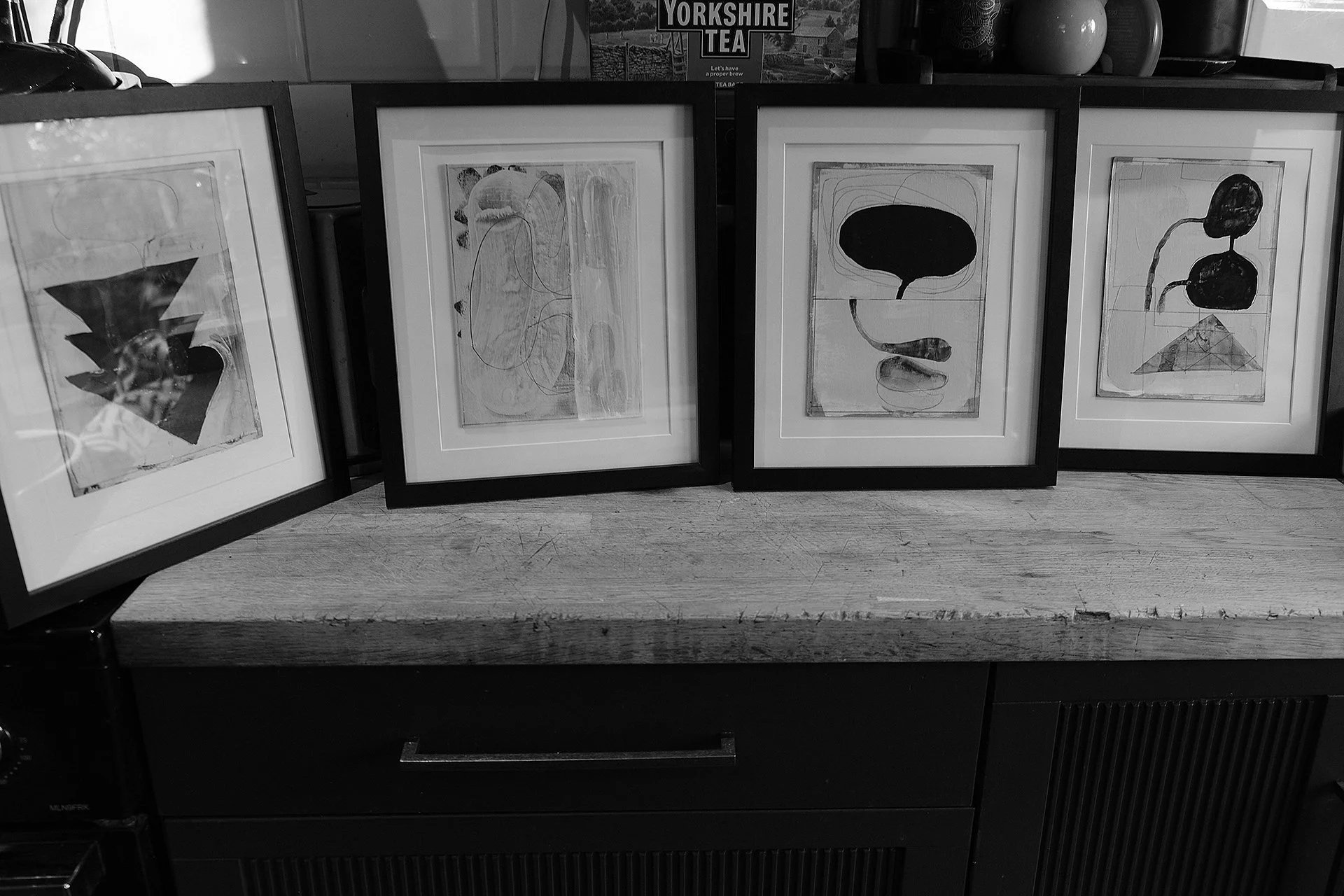 Four framed abstract art prints displayed on a wooden surface, with a coffee machine partially visible on the left and decorative vases on the right in a room.