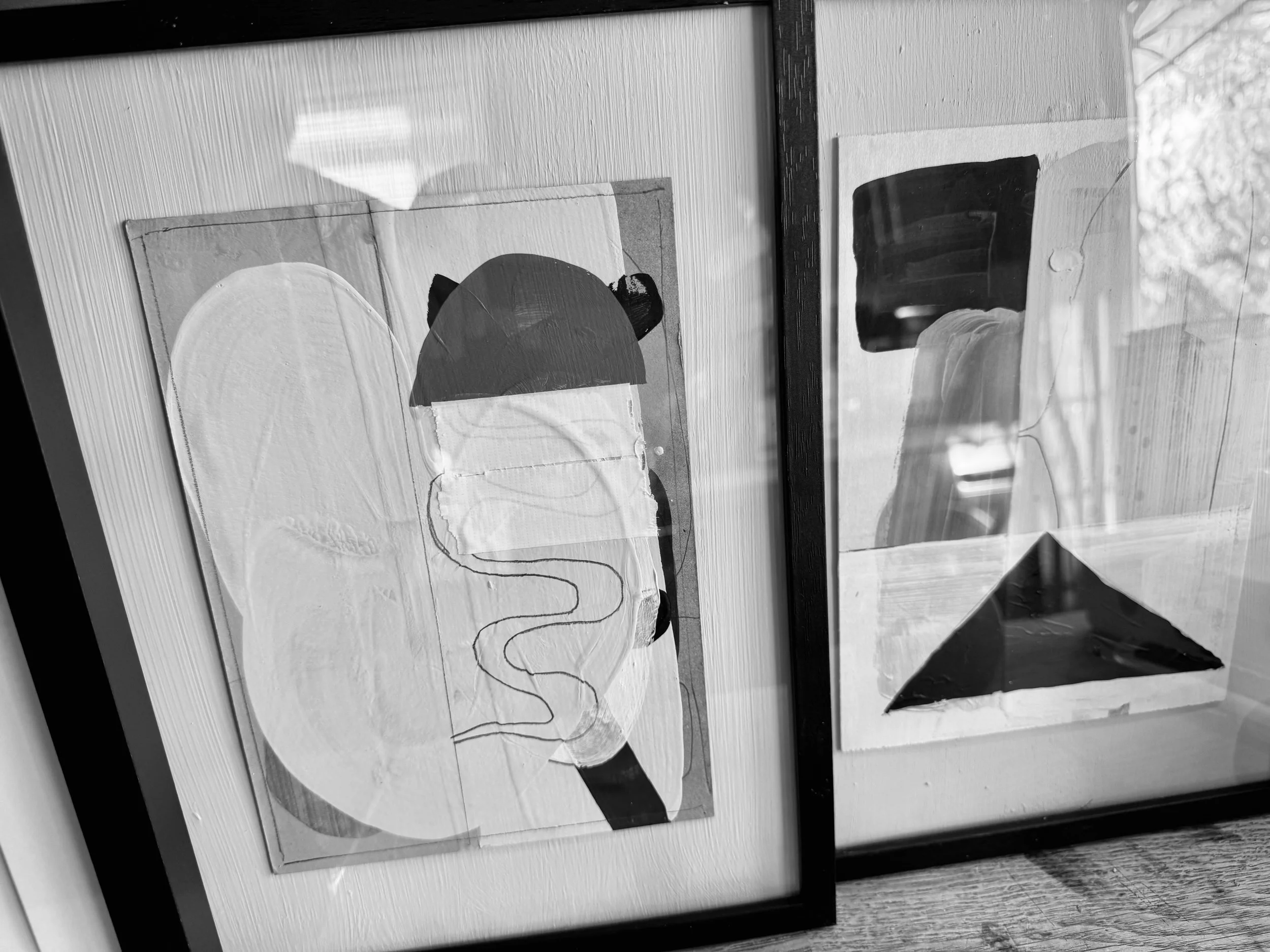 Two framed abstract art pieces leaning against a wall on a wooden surface. The left one features a stylized face with minimalistic features, including a white oval shape for the face, black hair with two rounded shapes resembling ears, and a continuo