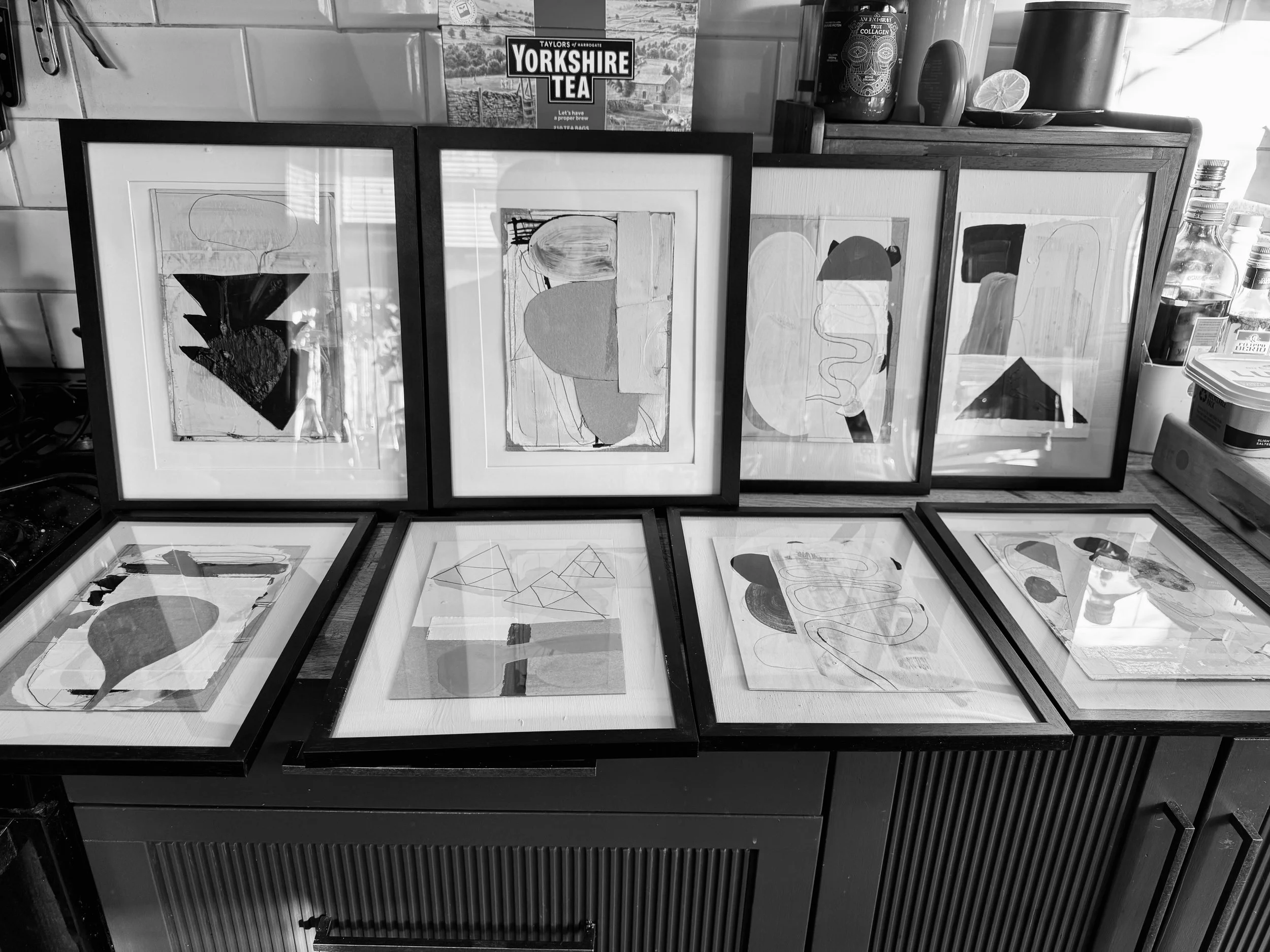 A display of abstract artwork in black and white, framed and arranged on a surface, with a box of Yorkshire Tea and various jars and containers visible in the background.