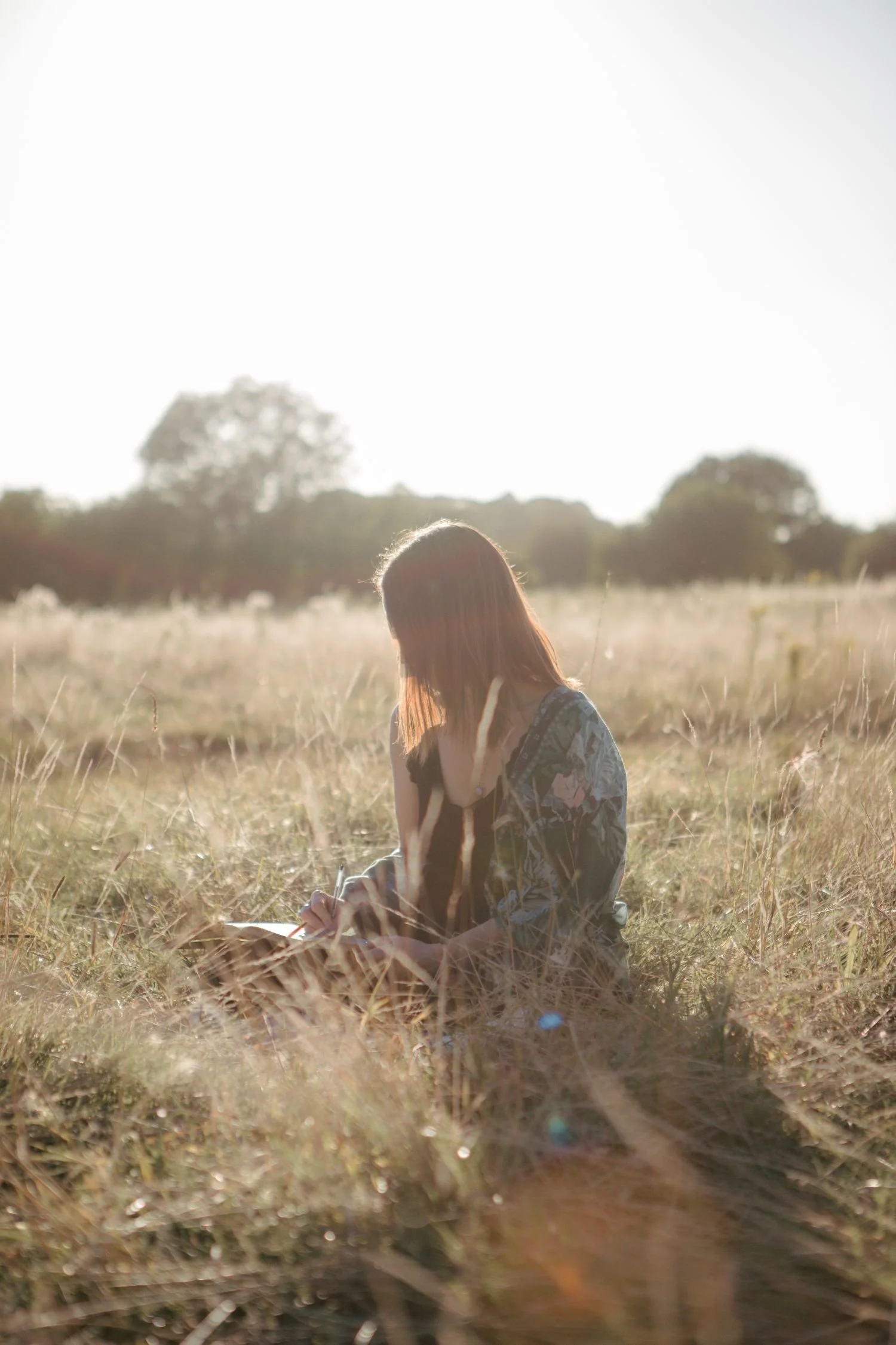 A woman sitting in a grassy field, writing or drawing in a notebook with sunlight shining from behind her.