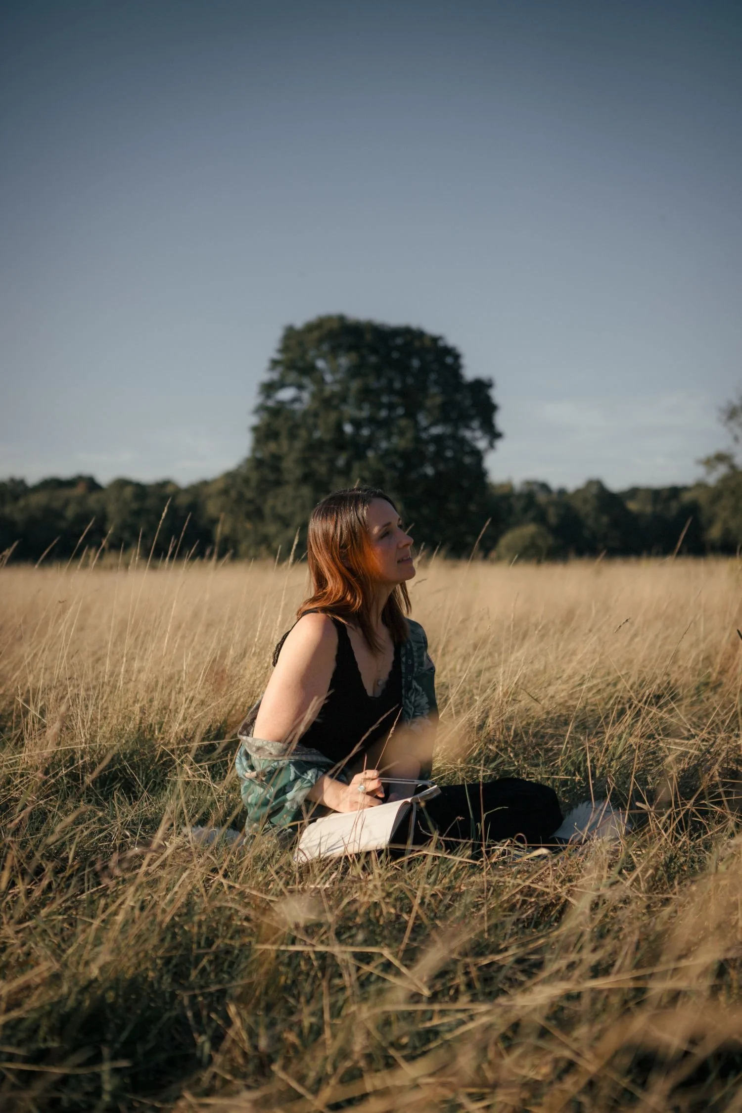 A woman sitting in a grassy field during daytime, holding a notebook and pen, with trees and a blue sky in the background.
