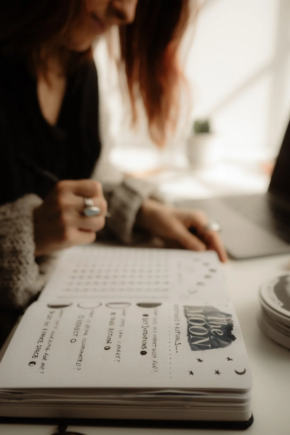Person sitting at a desk, writing in a notebook titled "the MOON," with a laptop and a stack of coasters nearby.