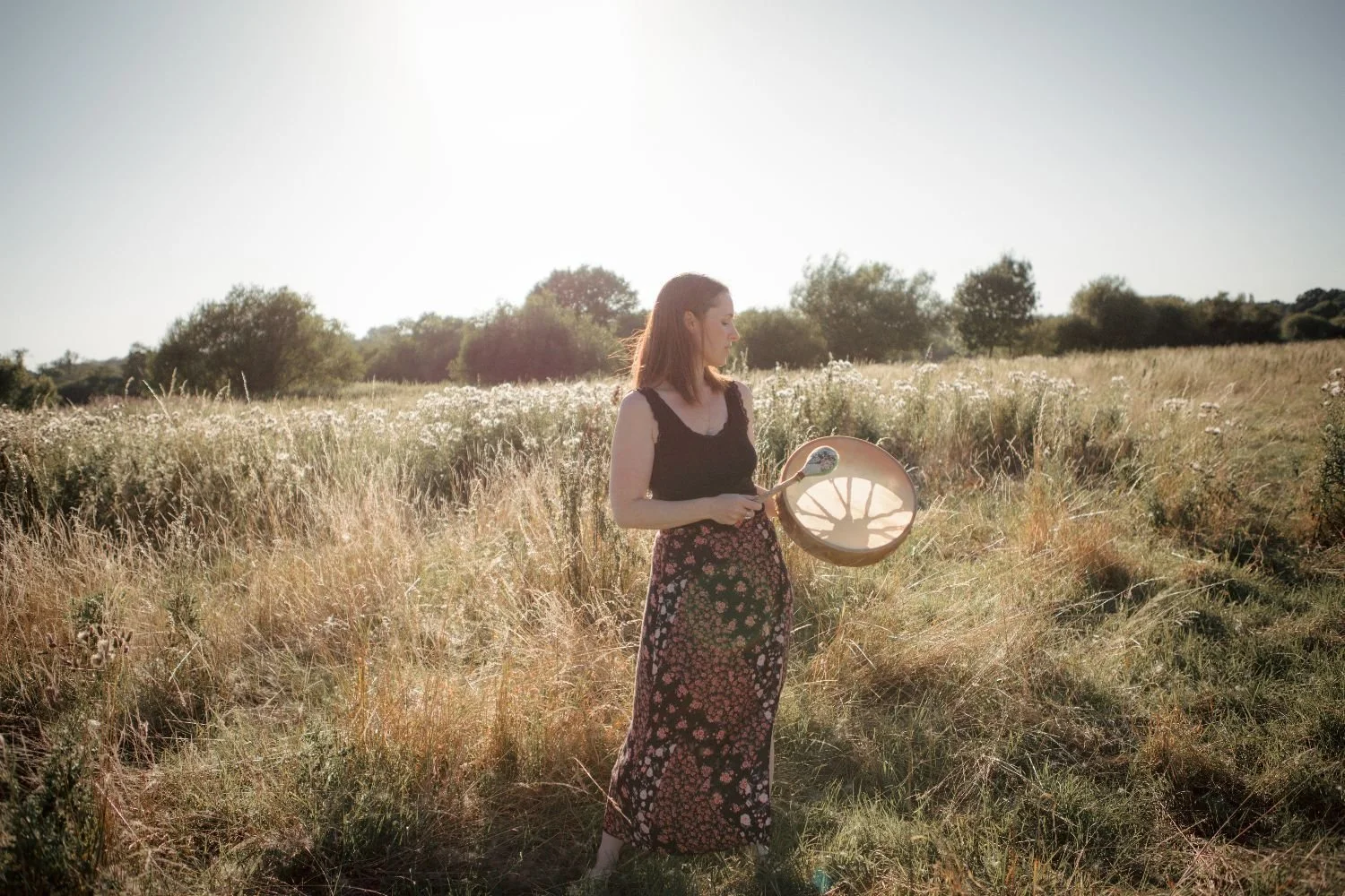 A woman standing in a grassy field holding a tambourine, with sunlight flooding the scene and trees in the background.
