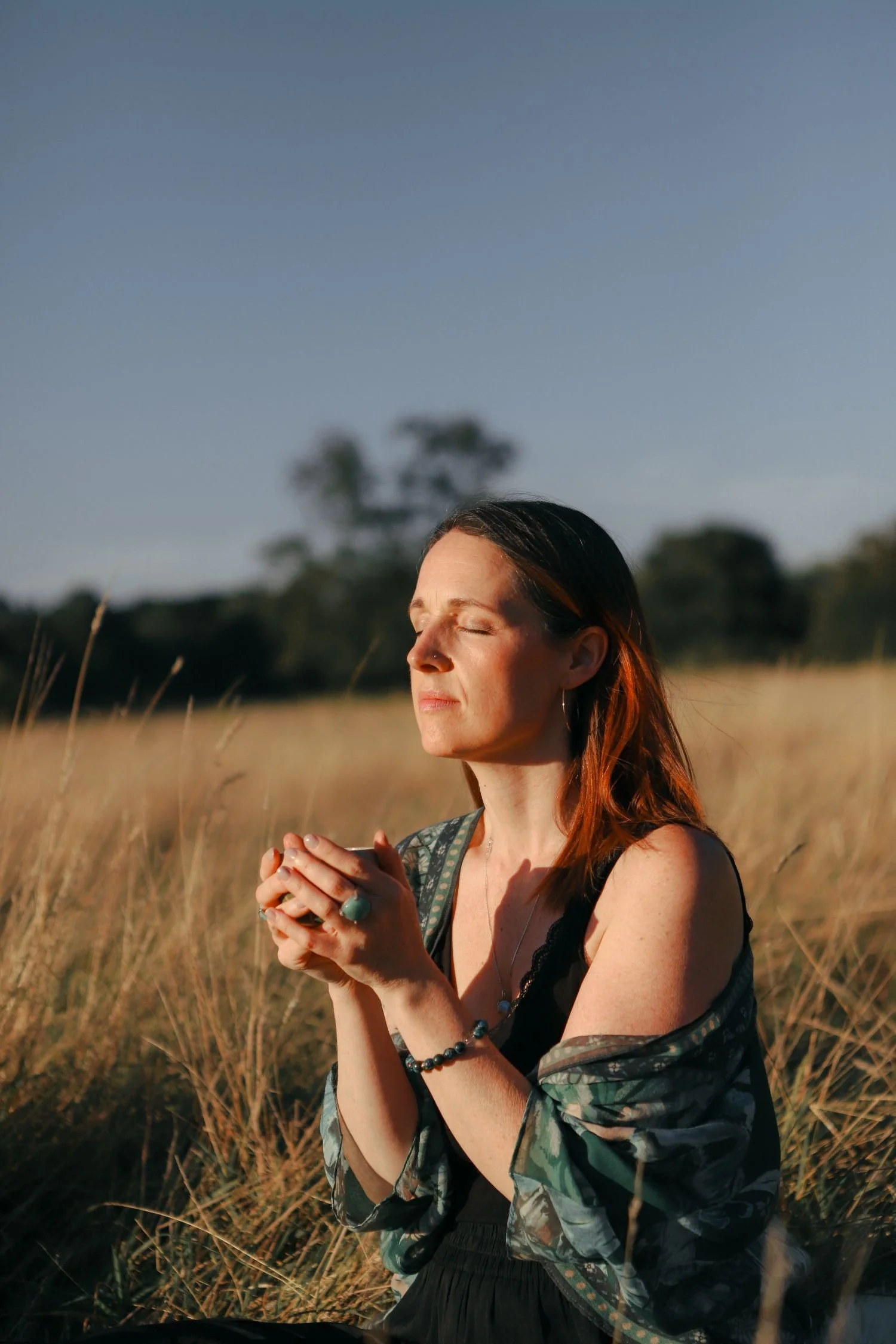 A woman in a black dress and floral shawl sitting outdoors in a field of tall grass with her eyes closed, holding a small object or cup with both hands, enjoying the sunlight during sunset.