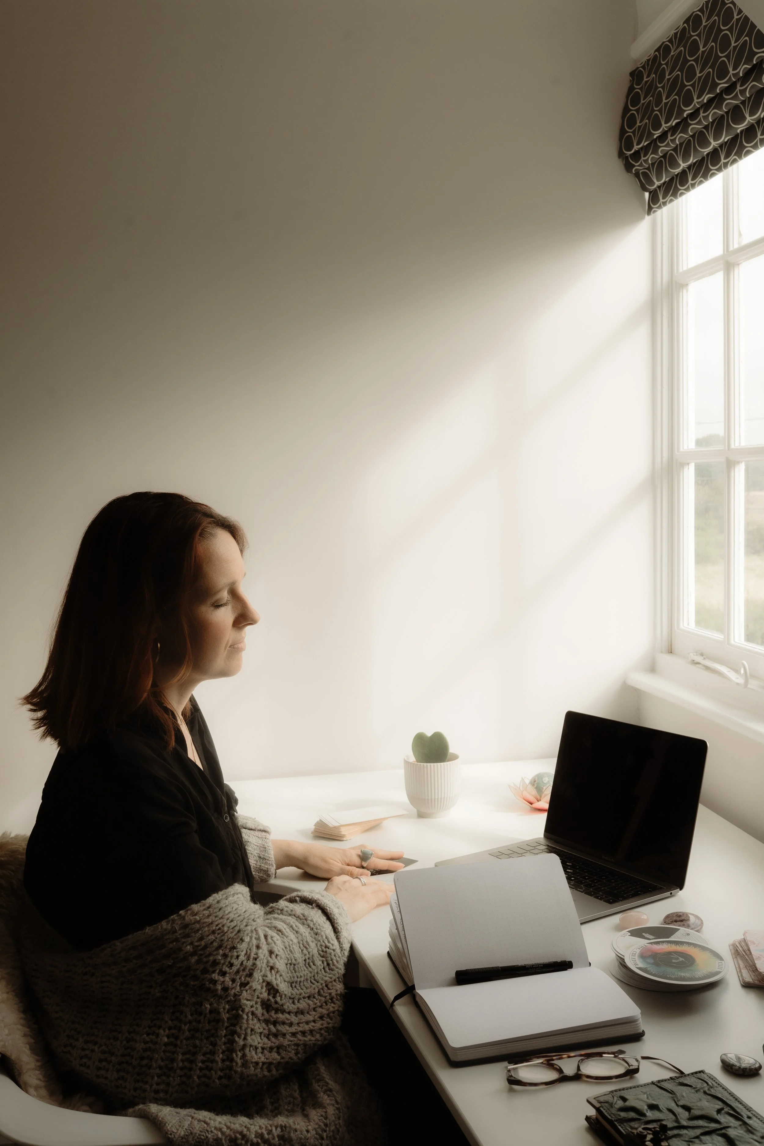 A woman with red hair and a gray knitted sweater sitting at a white desk with a laptop, open notebooks, and reading glasses, in a bright room with a large window, a small potted plant, and decorative items.