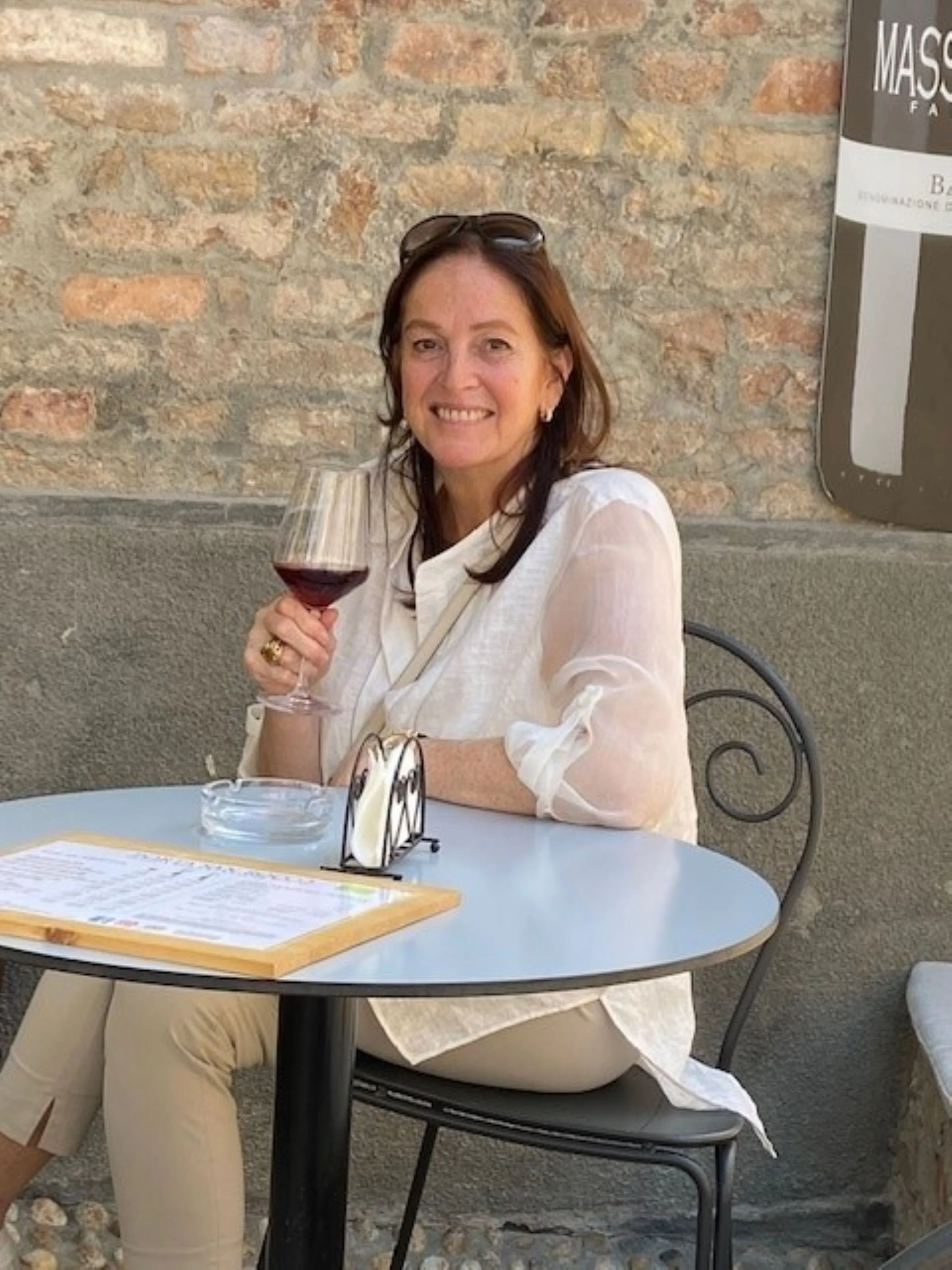 A woman with brown hair, wearing a white blouse and sunglasses on her head, sitting at a round table, smiling, and holding a glass of red wine in an indoor setting with a brick wall background.