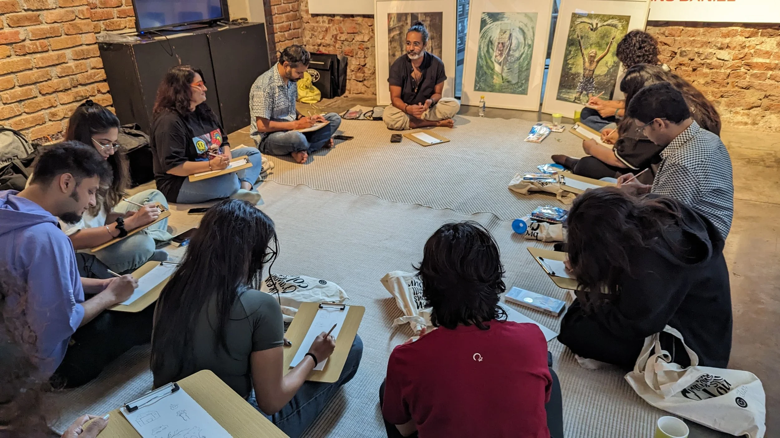 Group of people sitting on a carpeted floor in a circle, taking notes during a discussion or workshop in a room with brick walls and artwork.