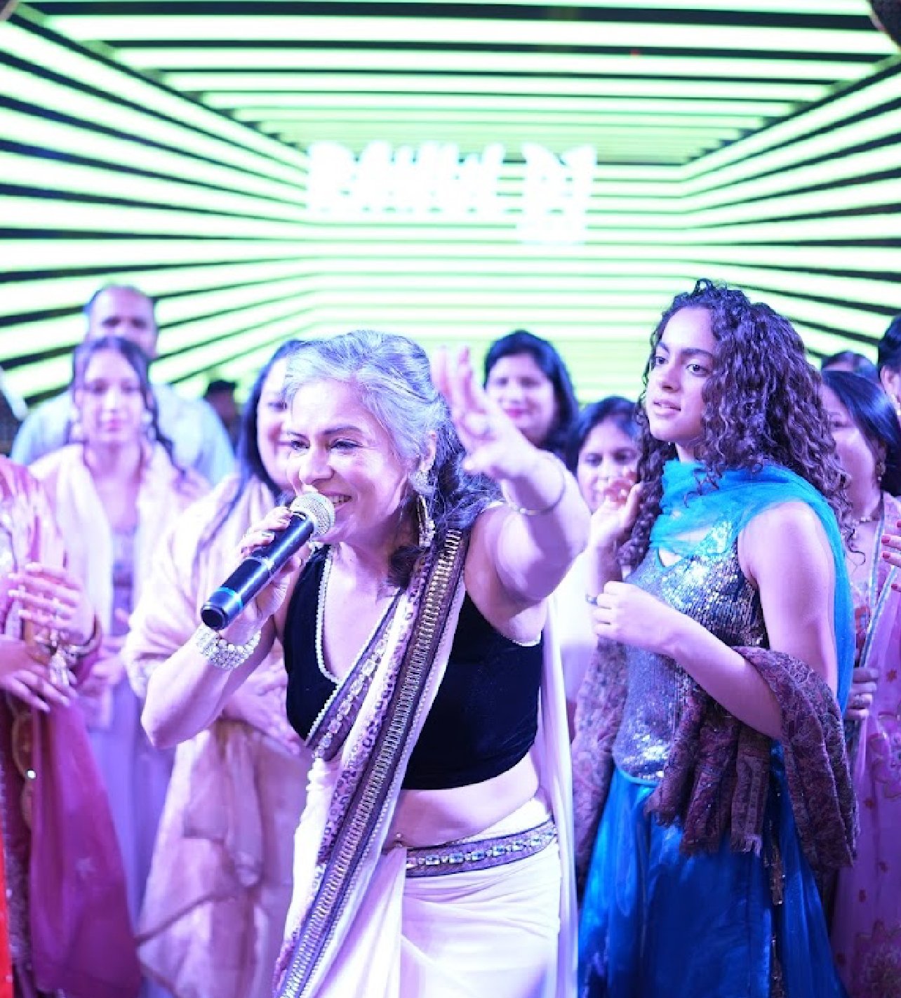 A woman singing into a microphone on stage with a group of people behind her, in front of a green and white striped LED background.