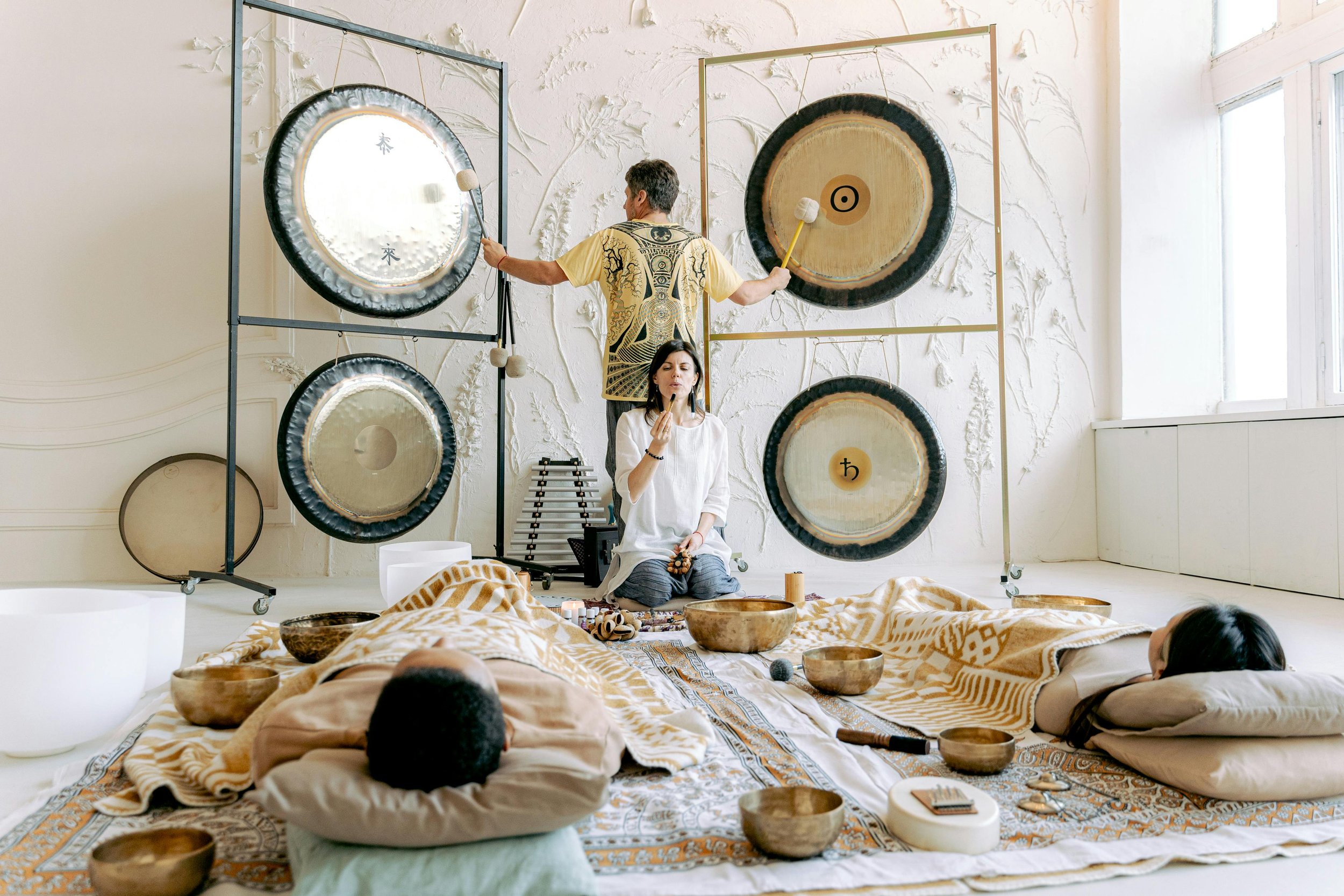 A group of people participate in a sound healing session inside a bright, softly-lit room. Two individuals lie on their sides on a colorful patterned rug, resting their heads on pillows, surrounded by singing bowls of various sizes. A woman seated cross-legged on the rug appears to be playing a small gong or singing bowl, with her eyes closed and hands in a meditative posture. Behind her, a man is standing and hitting a large gong mounted on a stand, with two other gongs behind him. The room has a textured white wall with artistic designs, and on the right side, there are large windows allowing natural light to fill the space.