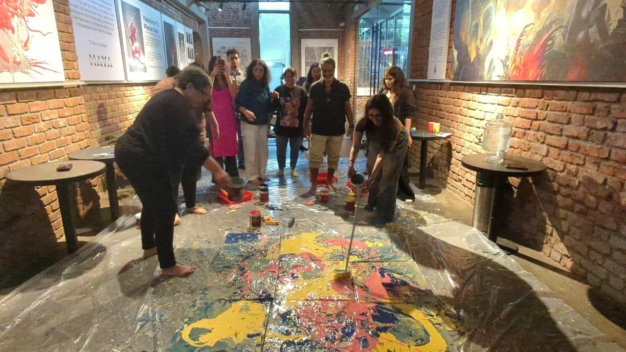 Group of people creating an abstract painting on a large canvas laid on the floor in a room with brick walls and artwork displayed on the walls.