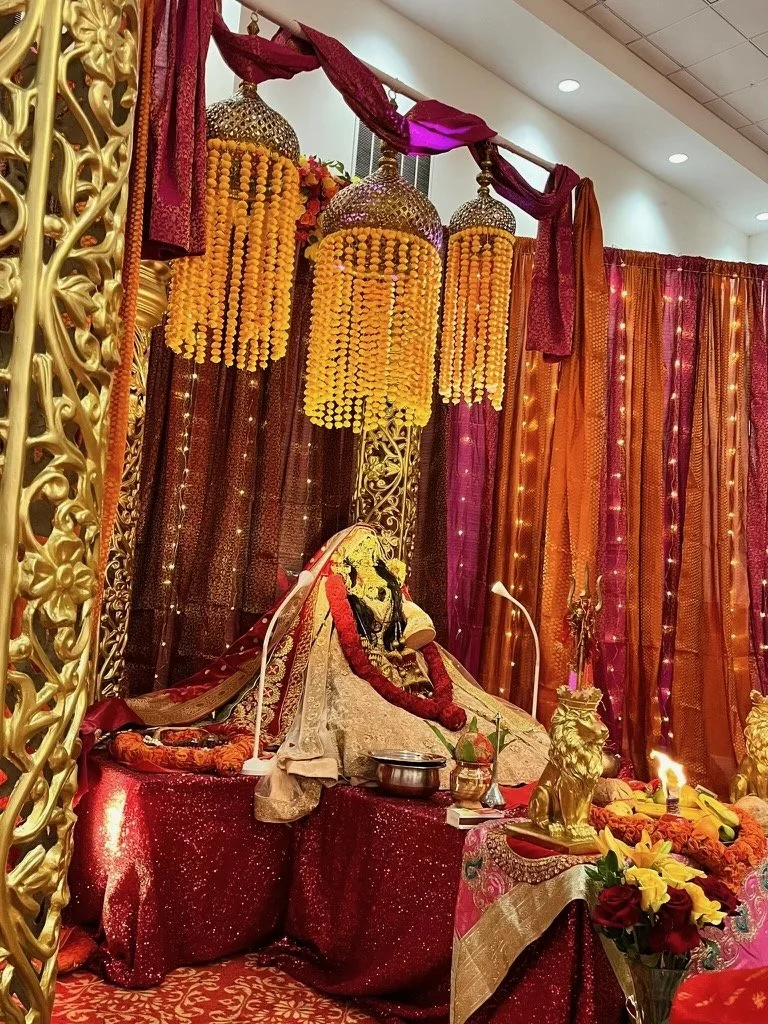 Hindu deity idol decorated with garlands, surrounded by ornate drapes, hanging lanterns, and floral arrangements, within a decorated prayer altar.