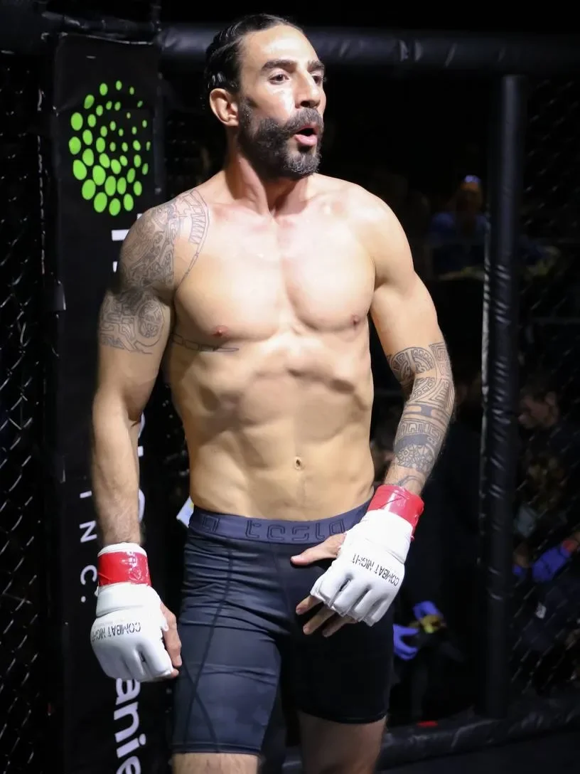 A shirtless male mixed martial artist with tattoos, wearing MMA gloves and shorts, standing inside an MMA cage with black mesh fencing, looking focused.
