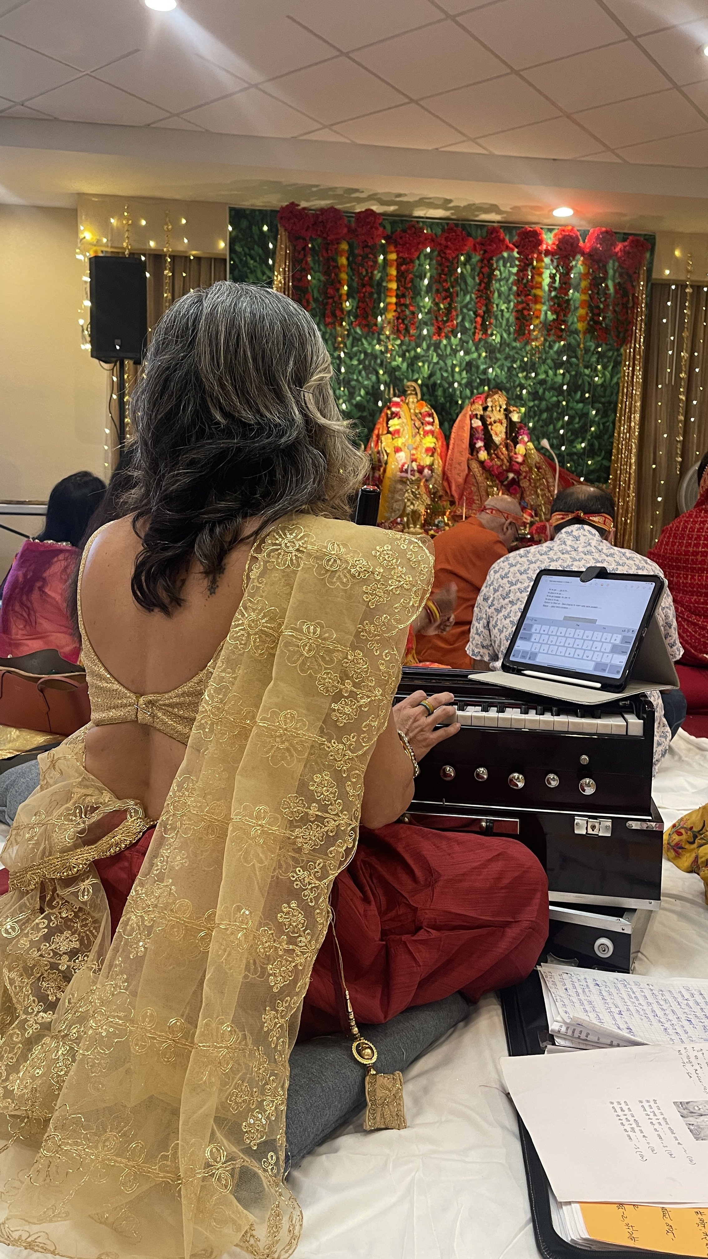 A woman dressed in traditional Indian attire, sitting on a man’s lap, playing a harmonium at a religious or cultural celebration with decorated idols and floral arrangements in the background.